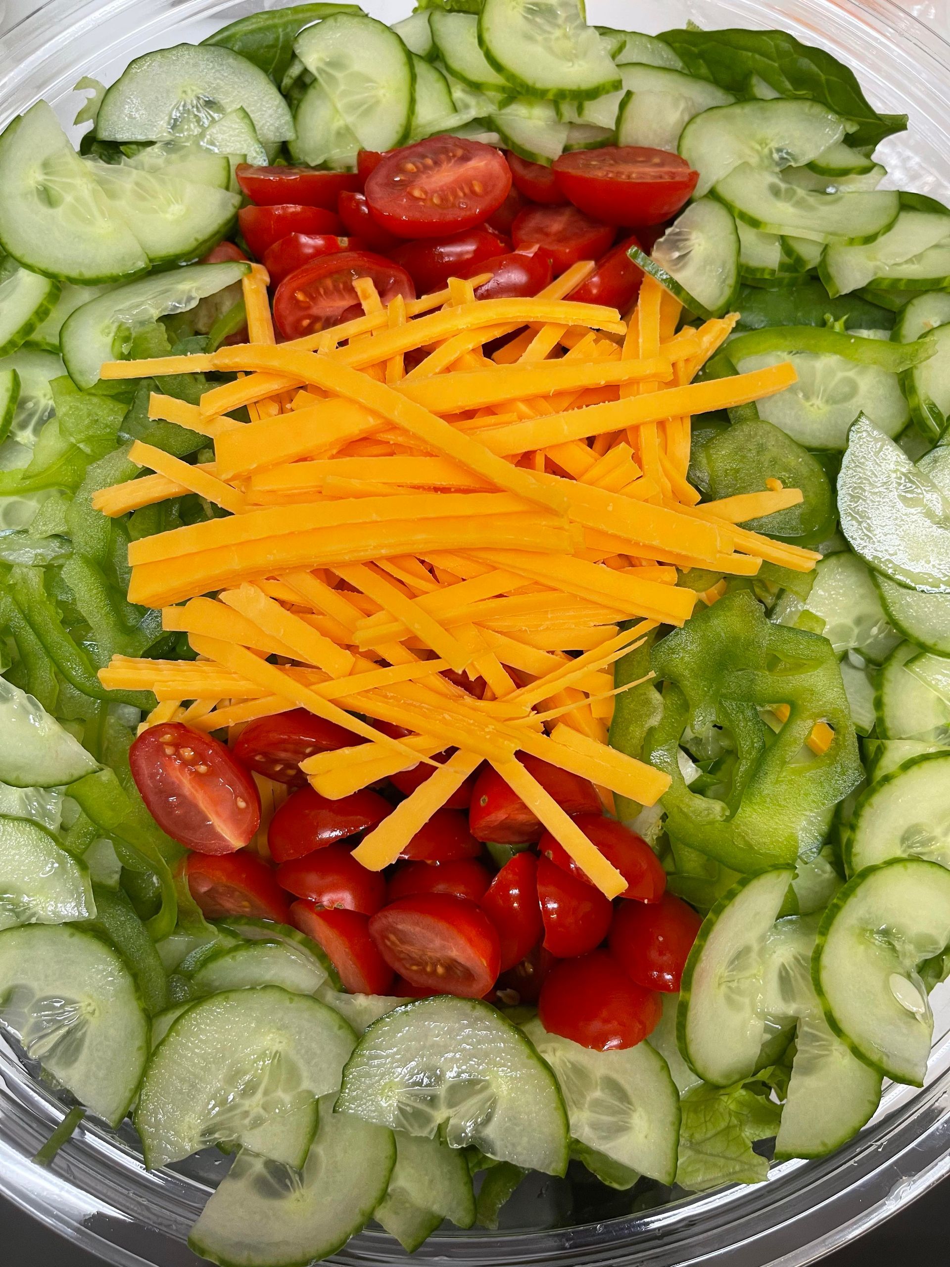 A salad in a clear bowl featuring sliced cucumbers, halved cherry tomatoes, and shredded orange cheddar cheese on top.