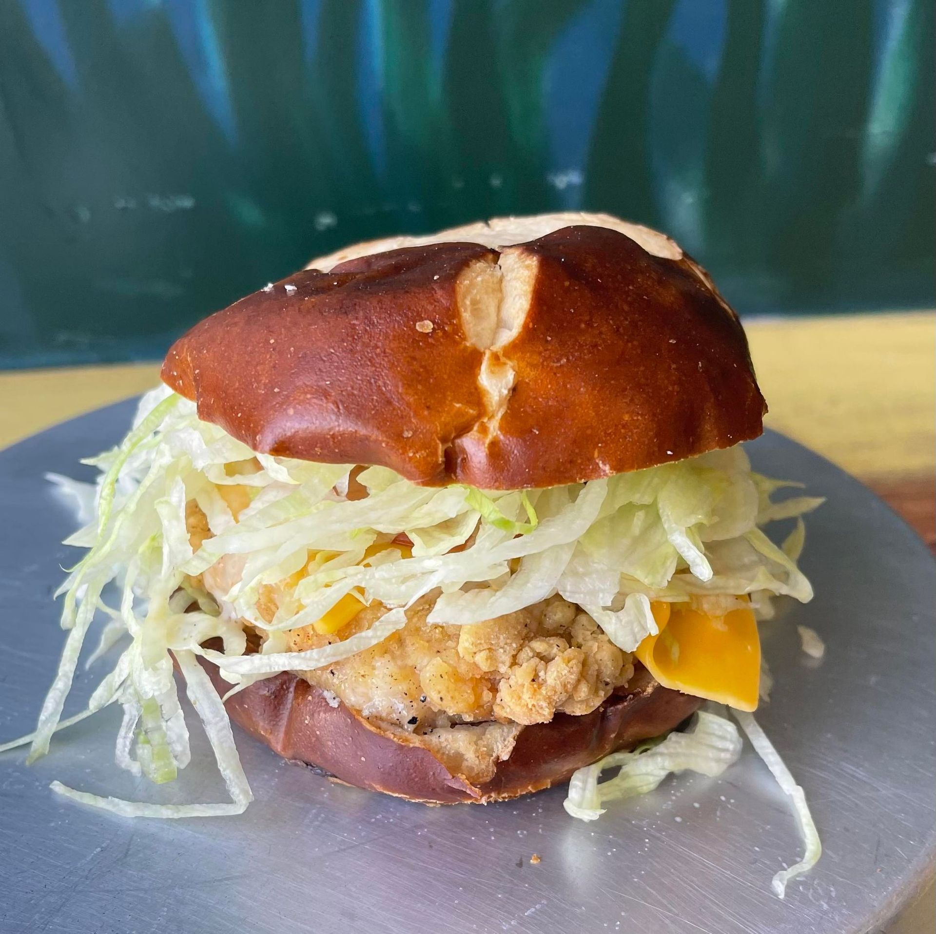 A breaded chicken sandwich with shredded lettuce and cheese on a pretzel bun, served on a metal plate.