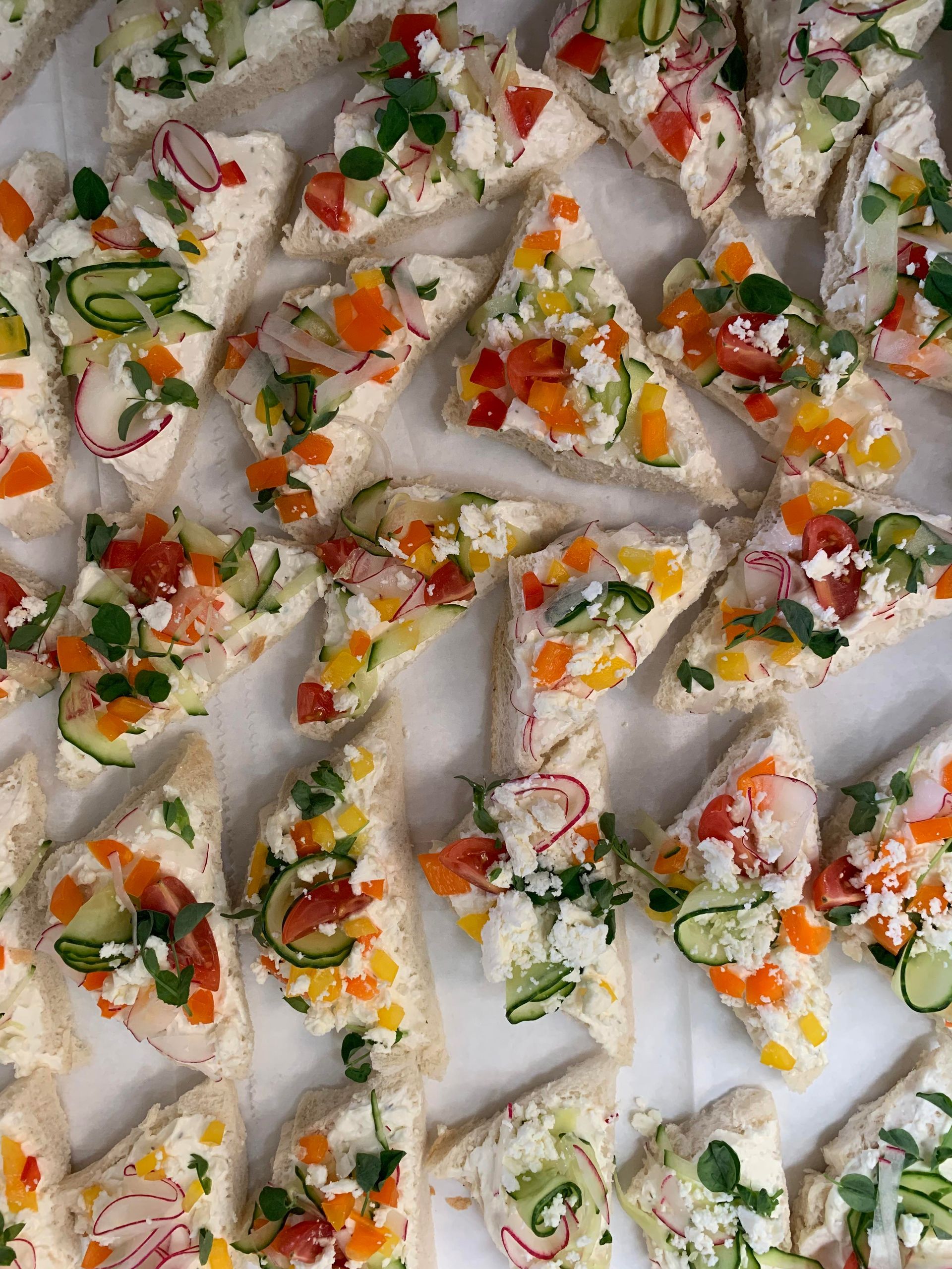 A tray of triangular sandwiches topped with creamy spread, diced bell peppers, cucumber slices, radishes, and fresh herbs.