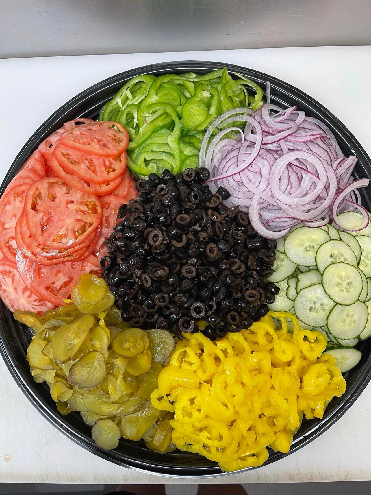 A circular party platter containing sections of sliced tomatoes, green peppers, red onions, cucumbers, olives, and pickles.