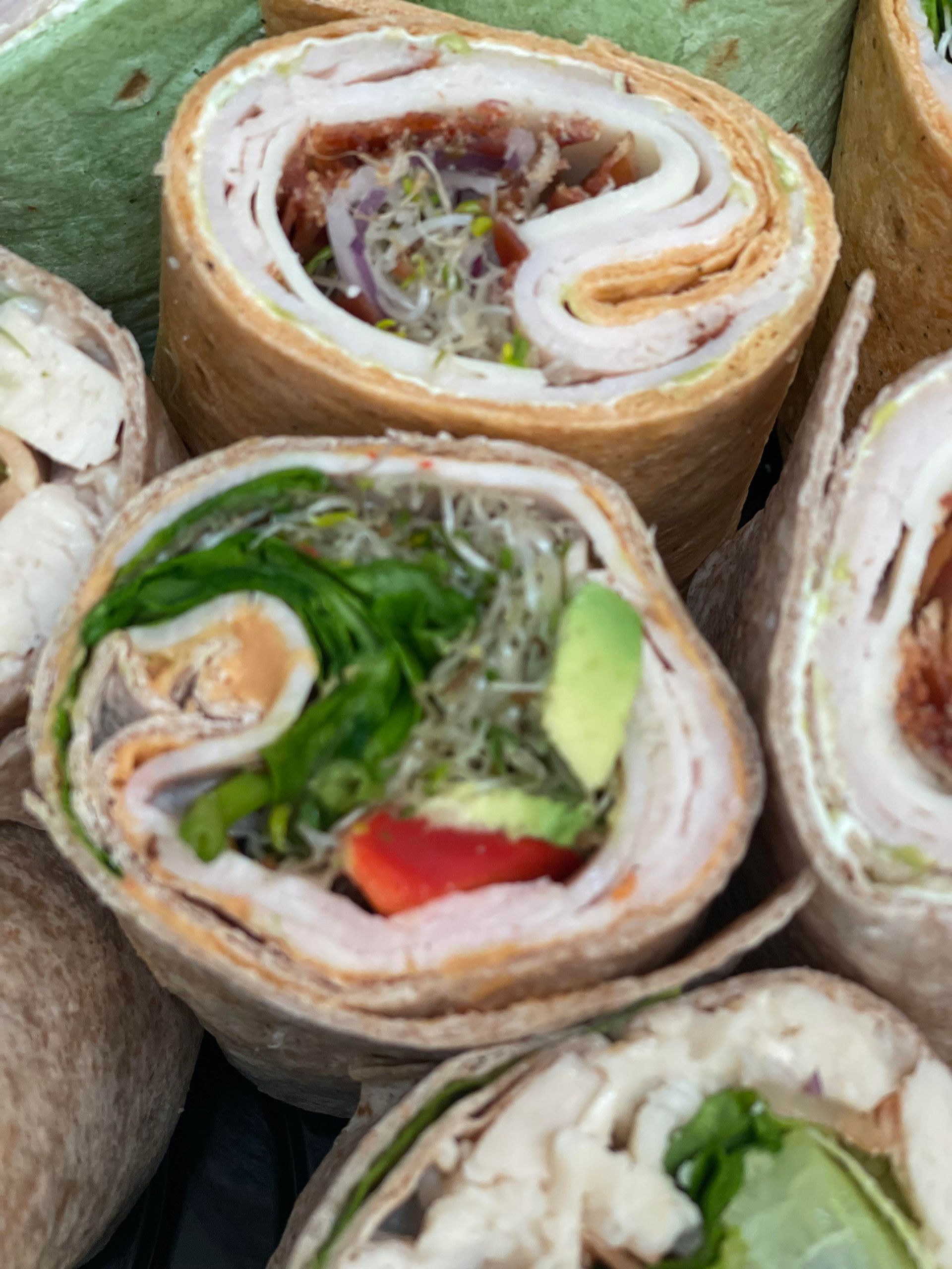 A close-up view of several sliced gourmet wraps filled with deli meat, avocado, sprouts, and fresh vegetables.