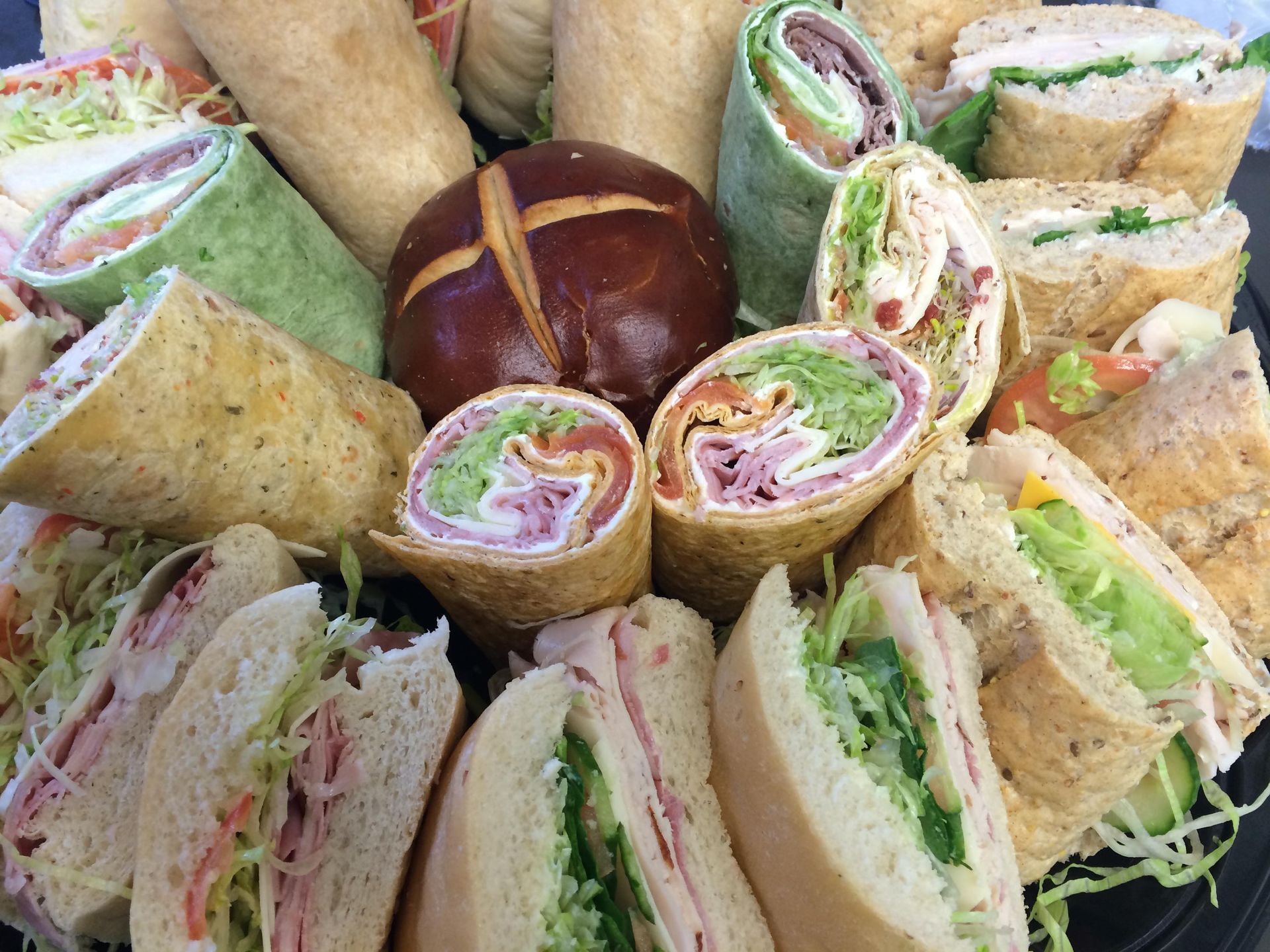 A party platter featuring an assortment of various deli sandwich wraps, sub sandwiches, and a central pretzel bun.