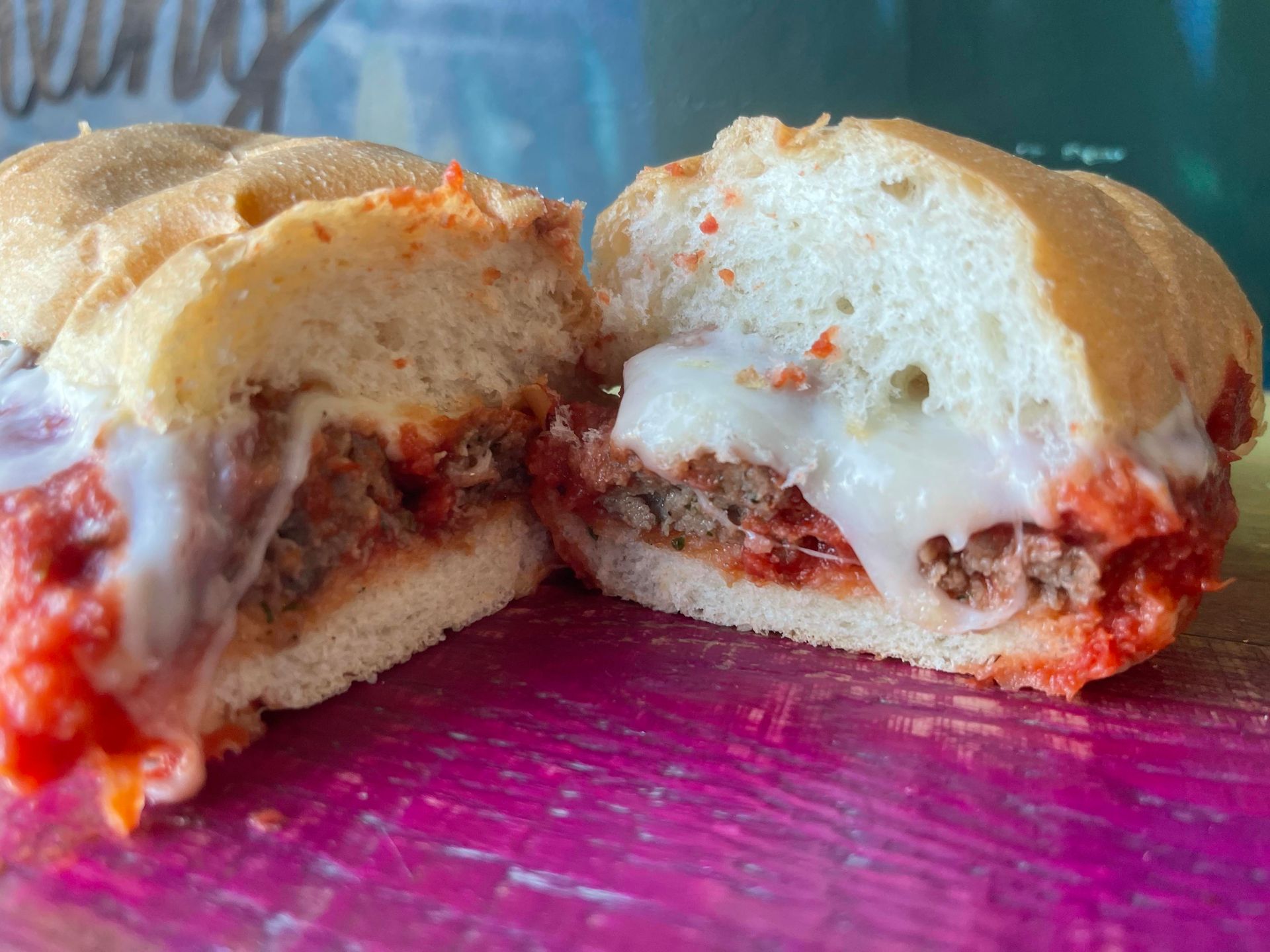 A halved meatball sub sandwich with melted white cheese and tomato sauce on a crusty roll, resting on a magenta surface.