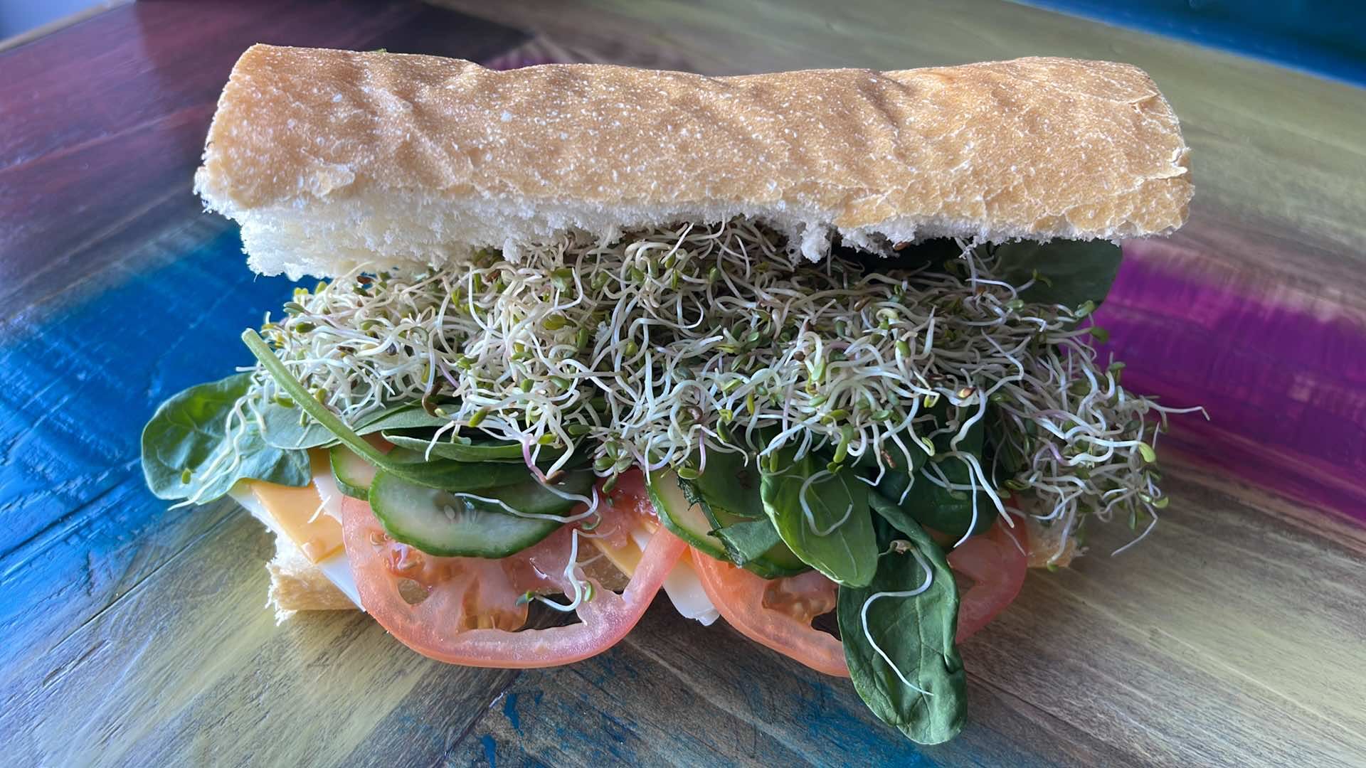 A vegetable sandwich with sprouts, spinach, tomato, and cucumber slices on a rustic baguette against a colorful background.