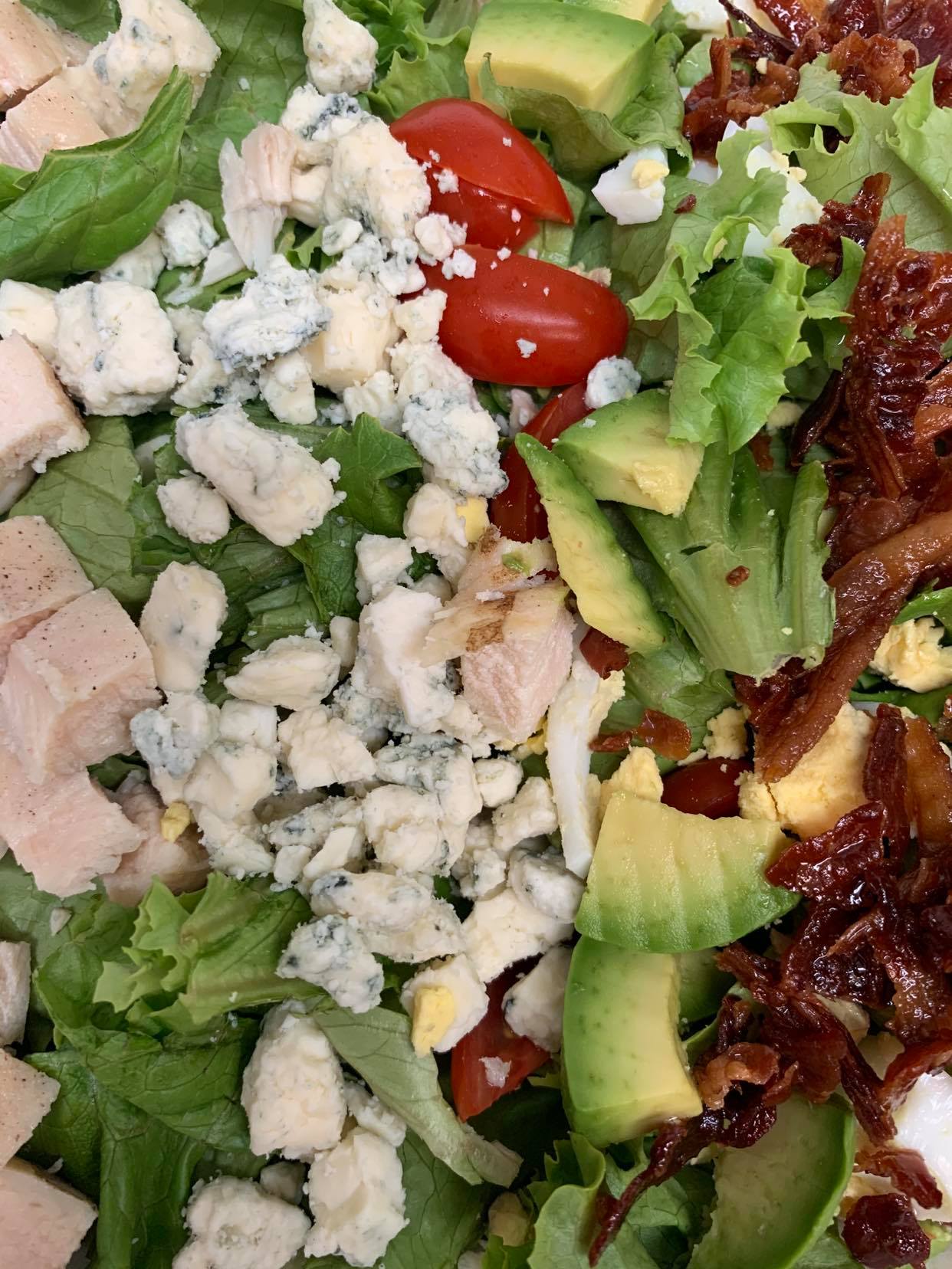 A Cobb salad featuring chopped chicken, blue cheese crumbles, sliced avocado, cherry tomatoes, and crispy bacon on greens.