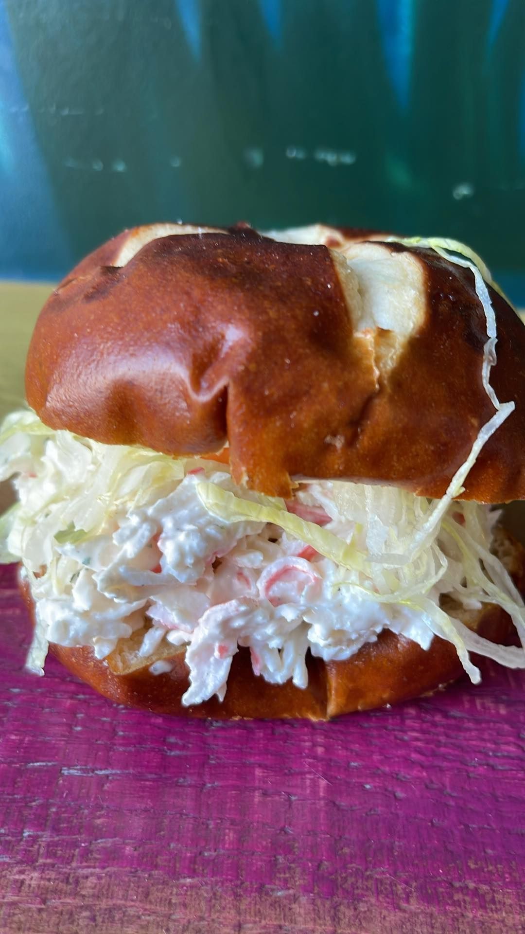 A crab salad sandwich with shredded lettuce on a toasted pretzel bun, sitting on a purple surface.