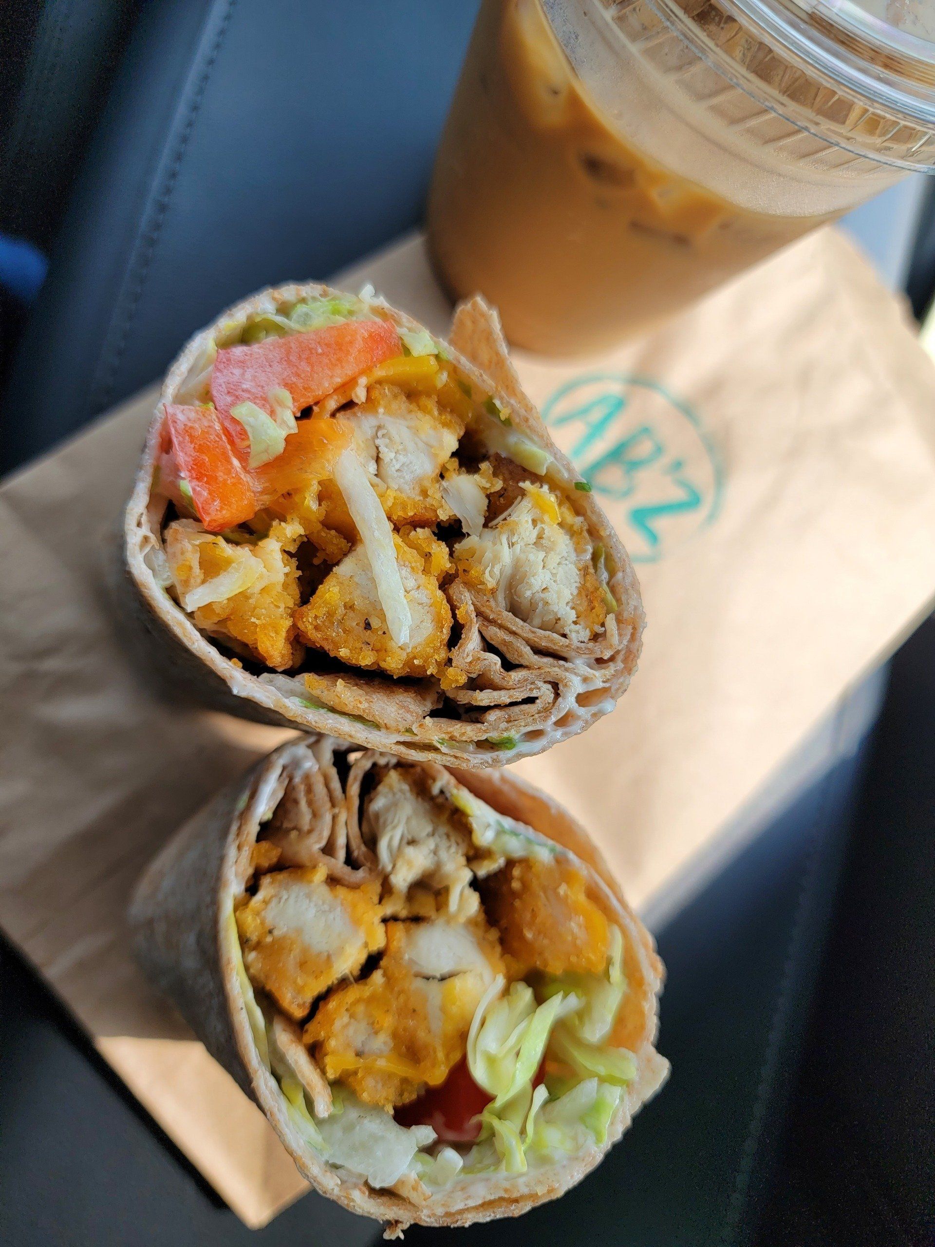 Two halves of a crispy chicken wrap with lettuce and tomatoes on a brown paper bag, next to an iced coffee.
