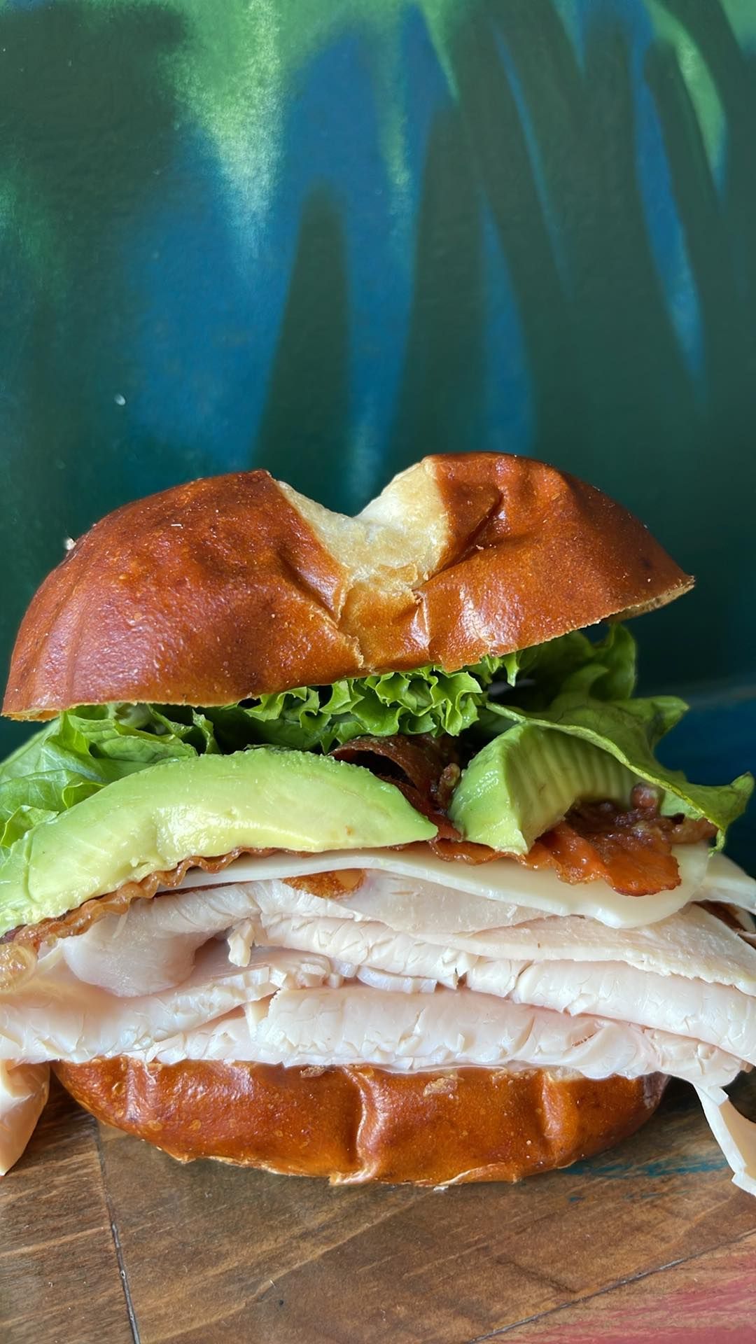 A turkey sandwich with sliced avocado, lettuce, and cheese on a pretzel bun, resting on a wooden surface.