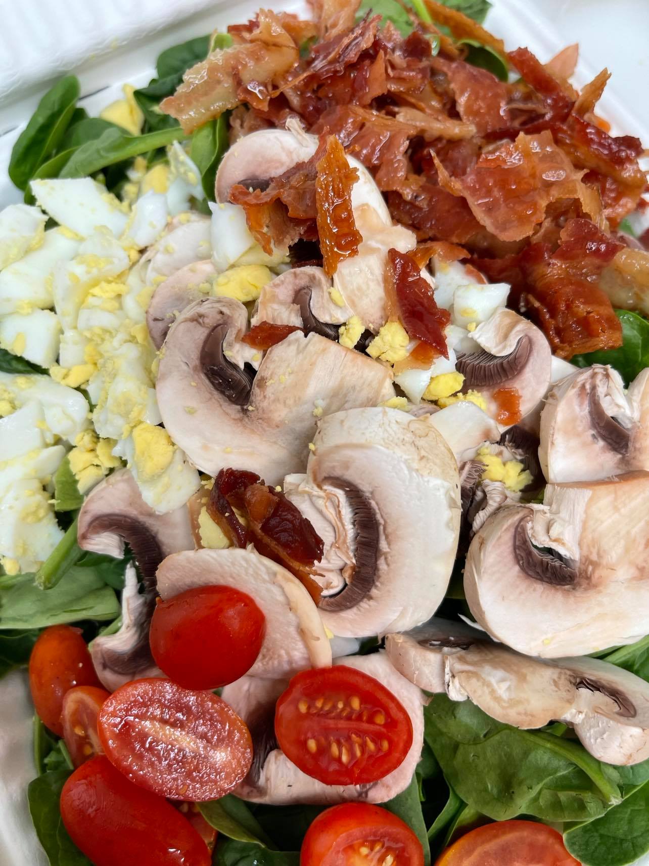 A salad featuring spinach, sliced mushrooms, cherry tomatoes, chopped hard-boiled eggs, and crispy bacon.
