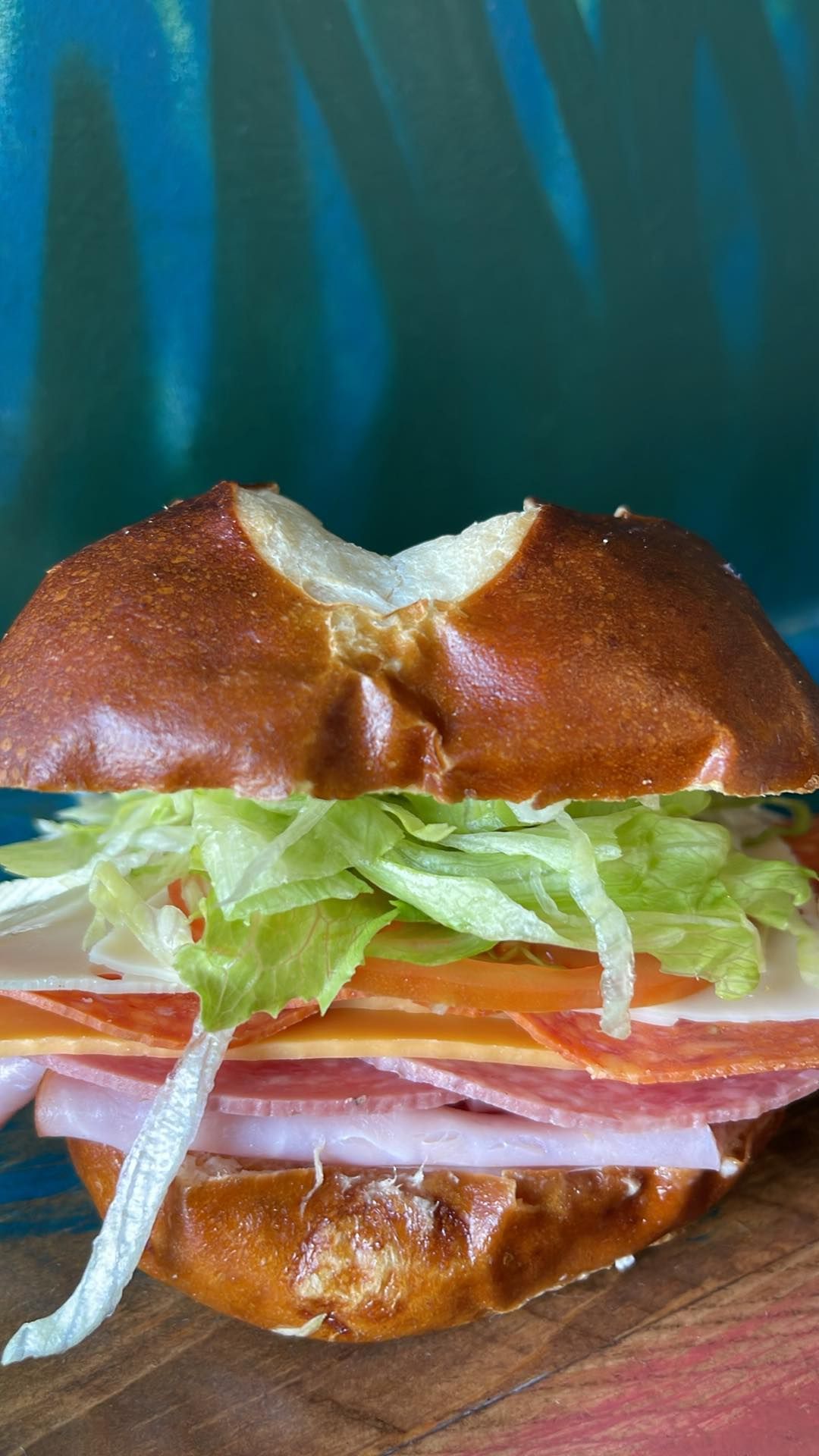 A deli sandwich with various meats, cheese, and shredded lettuce on a split pretzel bun, set against a teal background.