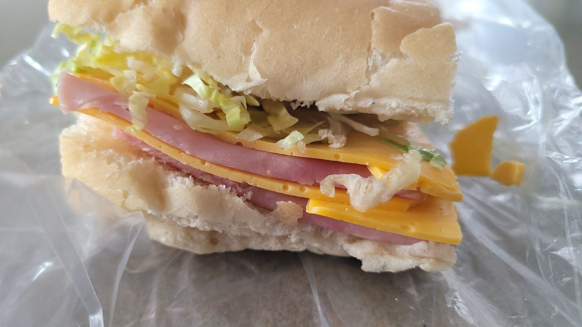 A ham and cheese sandwich on a soft white roll with shredded lettuce, sitting on a piece of clear plastic wrap.