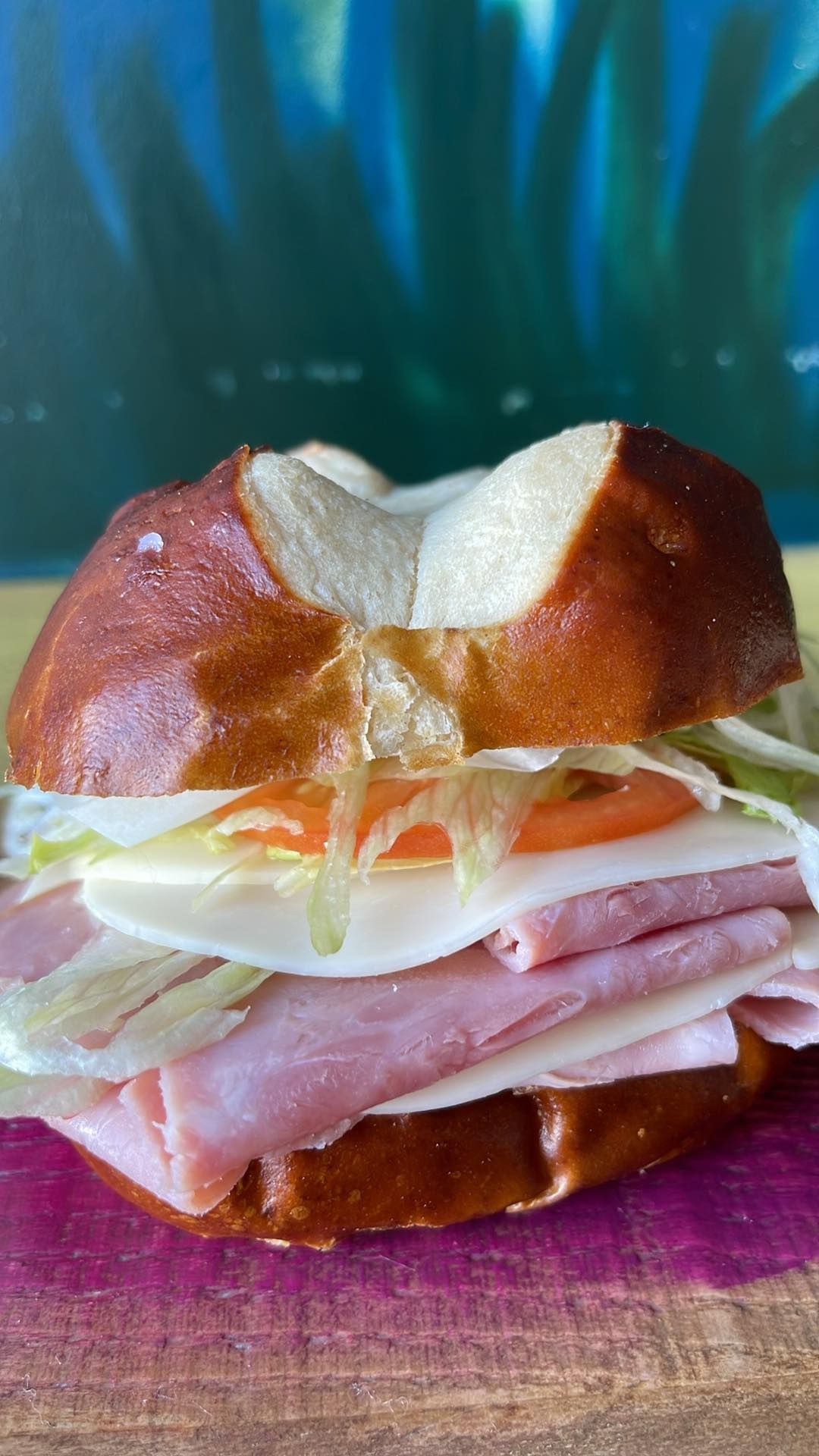 A ham and cheese sandwich on a pretzel bun with lettuce and tomato, resting on a wooden board against a blue background.