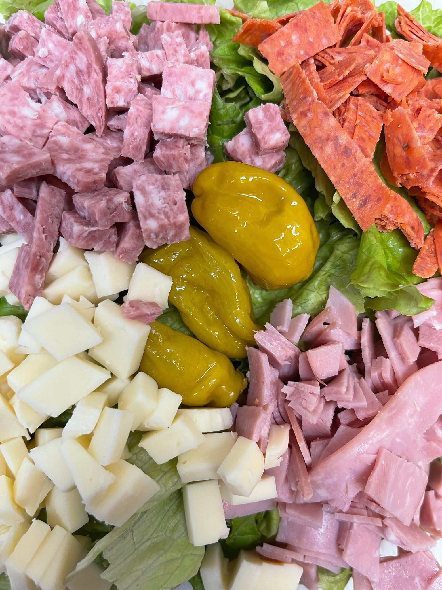 A salad bowl featuring diced salami, cubed cheese, sliced ham, pepperoni strips, and two yellow pepperoncini over lettuce.