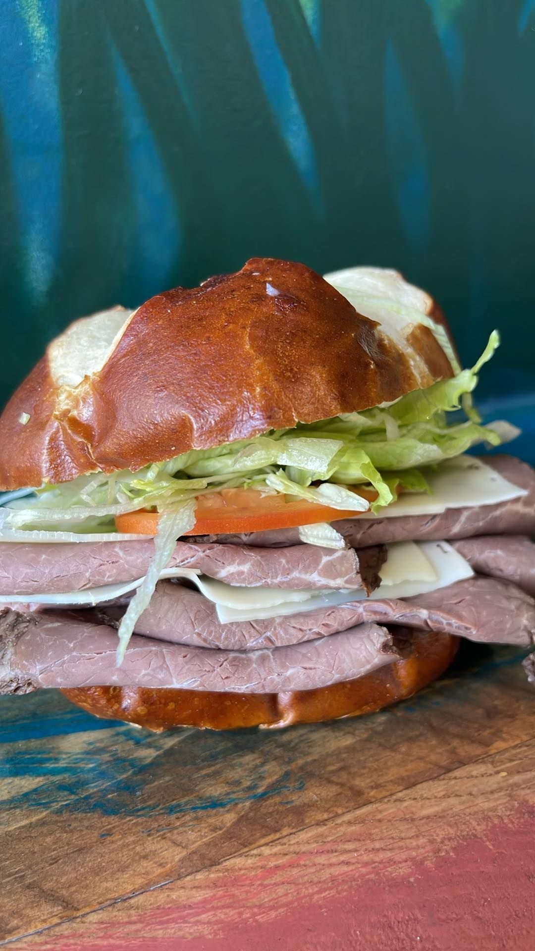 A roast beef sandwich with lettuce, tomato, and Swiss cheese on a dark pretzel bun, sitting on a rustic wooden board.