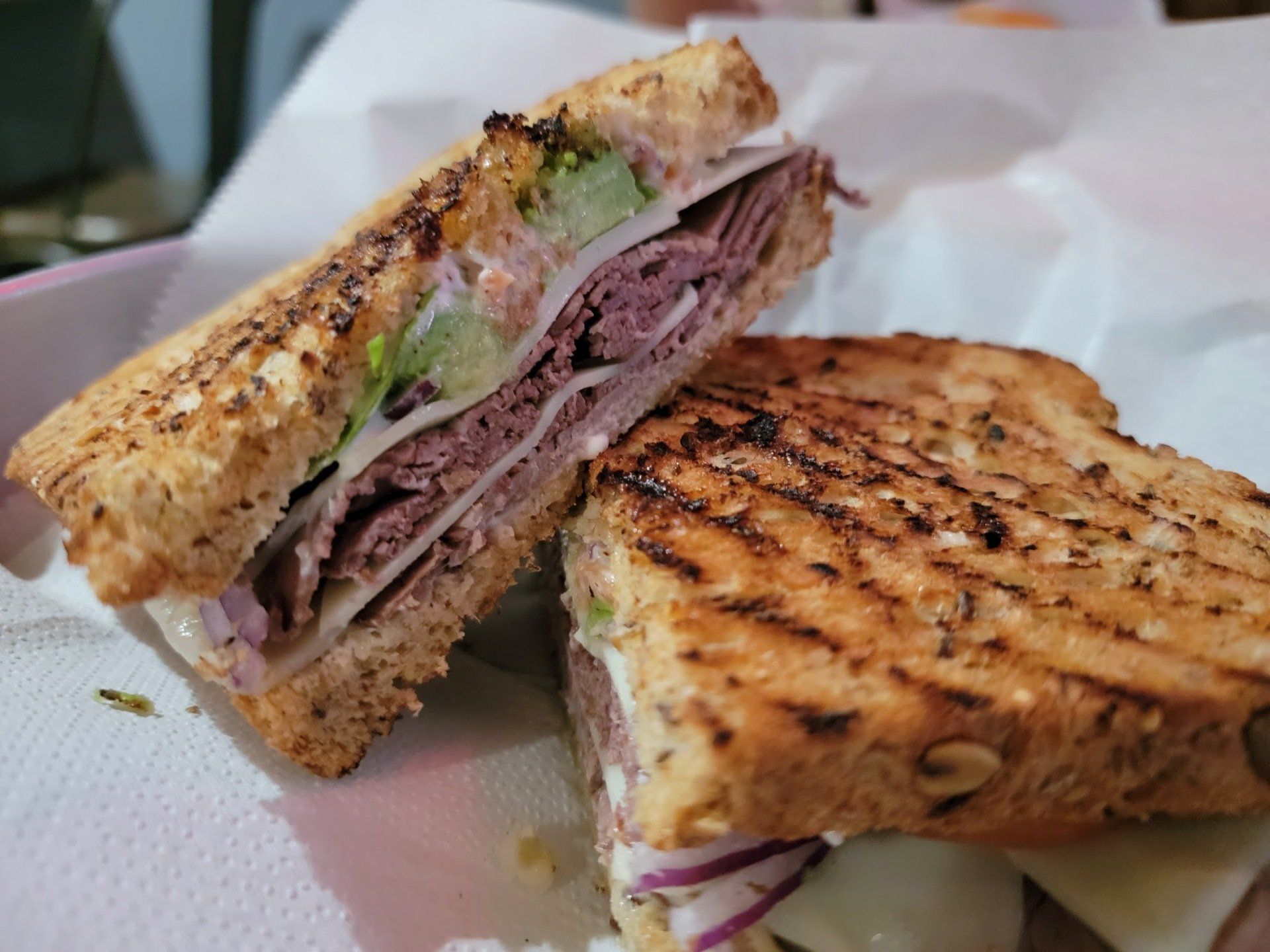 Two halves of a toasted deli sandwich with meat, cheese, lettuce, and onions on seeded bread, served on parchment paper.
