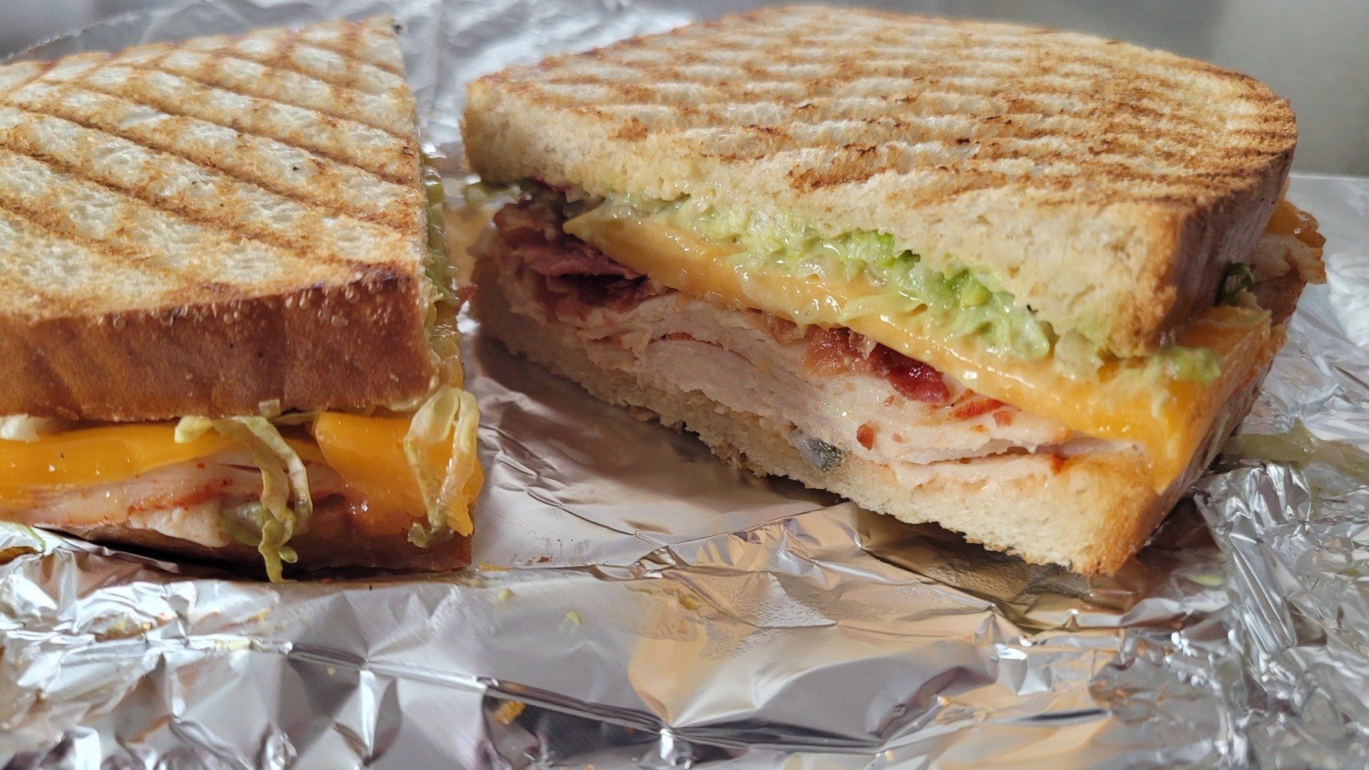 A grilled panini sandwich with melted cheese, turkey, and lettuce on toasted bread, sitting on a sheet of aluminum foil.