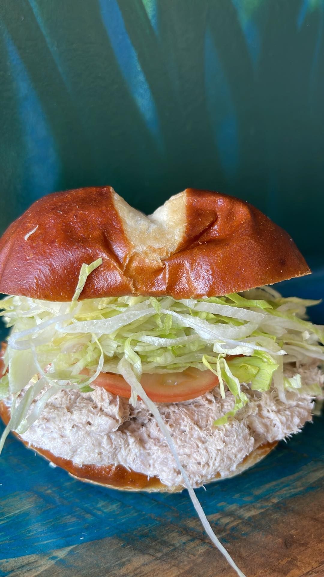 A tuna salad sandwich on a toasted pretzel bun with shredded lettuce and a tomato slice, set against a blue background.