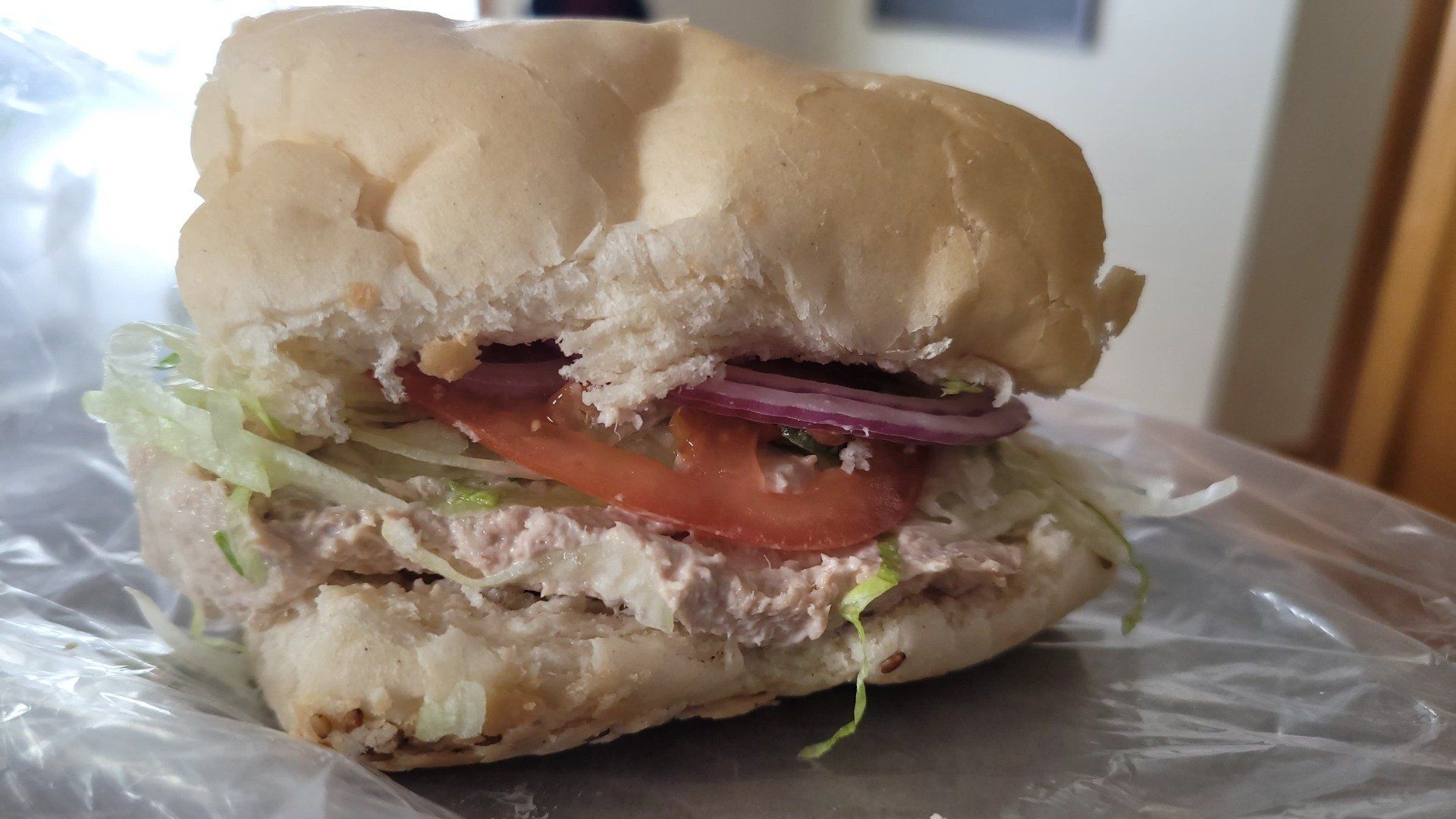 A tuna sandwich on a bun with lettuce, a slice of tomato, and red onion, resting on plastic wrap.