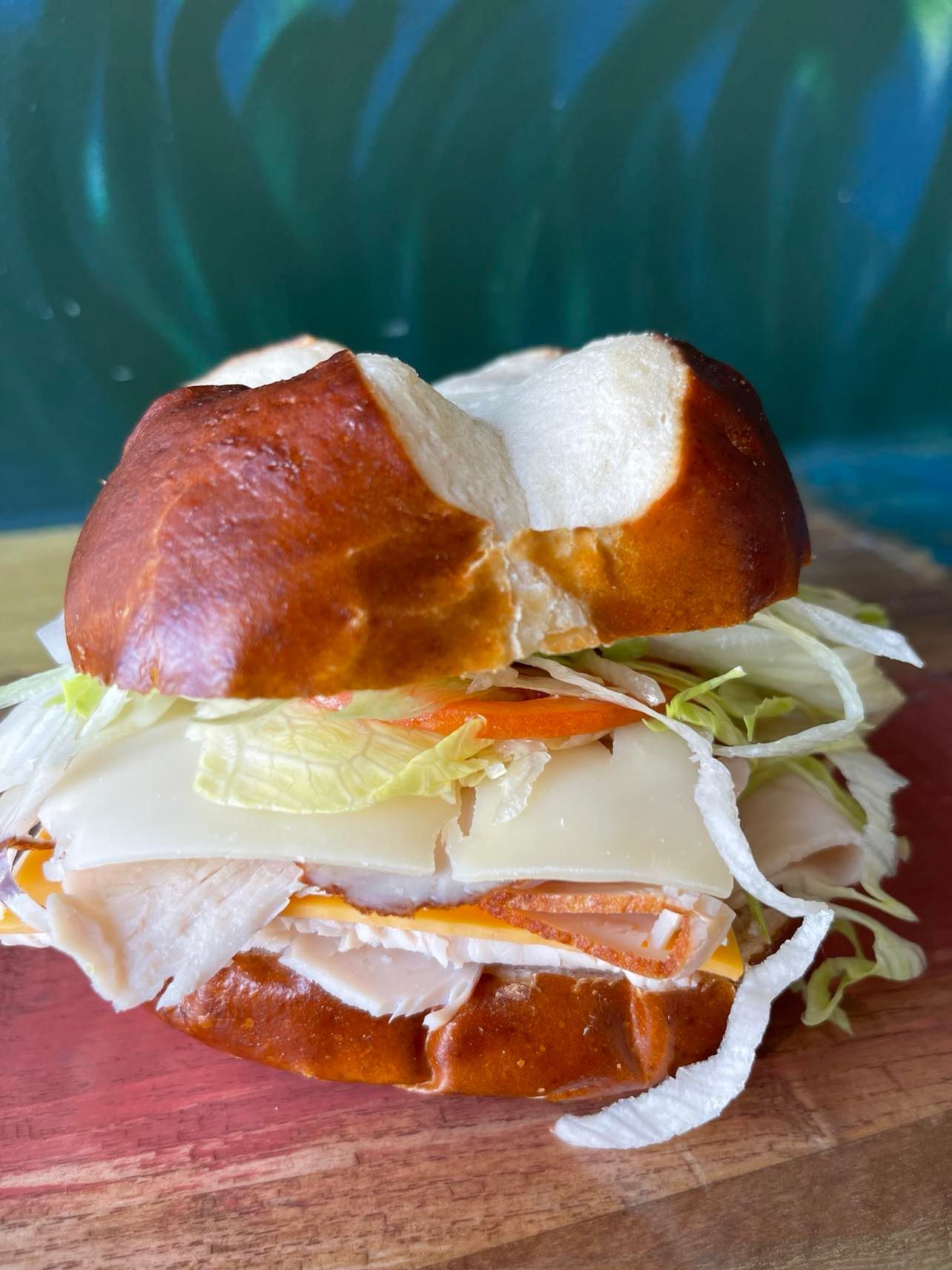 A turkey and cheese sandwich on a pretzel bun topped with shredded lettuce, served on a wooden board against a blue wall.
