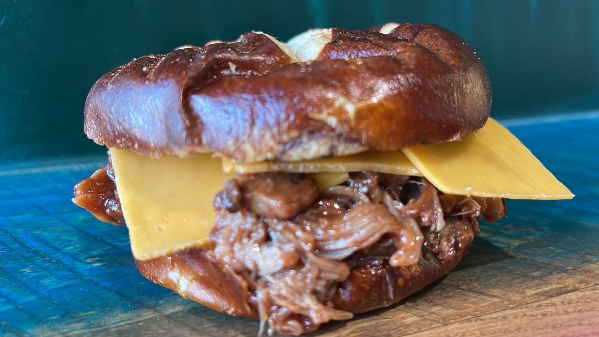 A pulled pork sandwich with slices of yellow cheese on a dark pretzel bun, resting on a wooden surface.