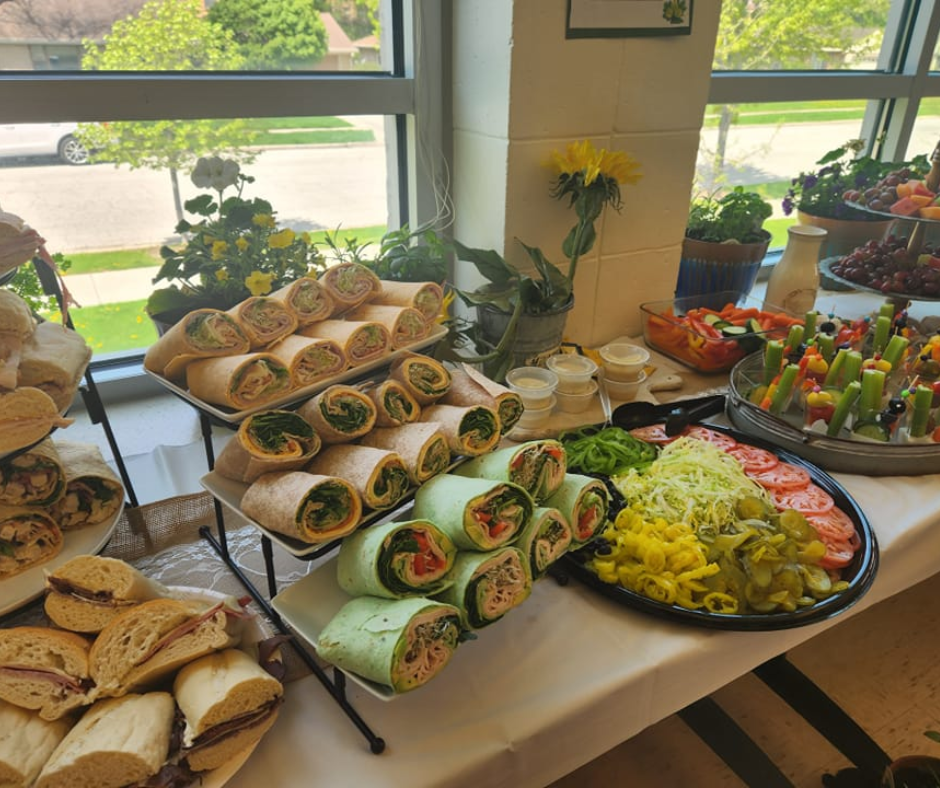 A buffet table with assorted wraps, sandwiches, a vegetable platter, and condiments near a sunny window.