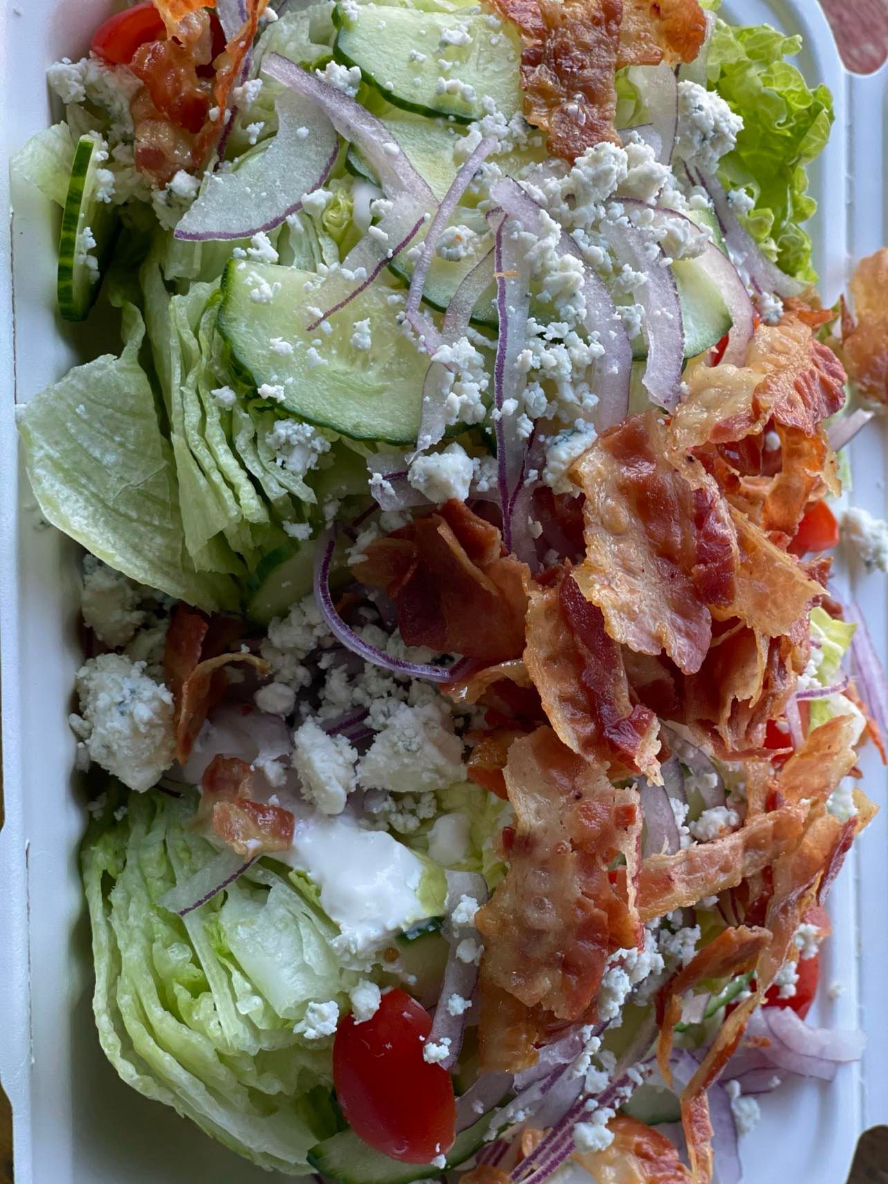 A wedge salad in a takeout container topped with bacon, crumbled blue cheese, red onions, cucumbers, and cherry tomatoes.