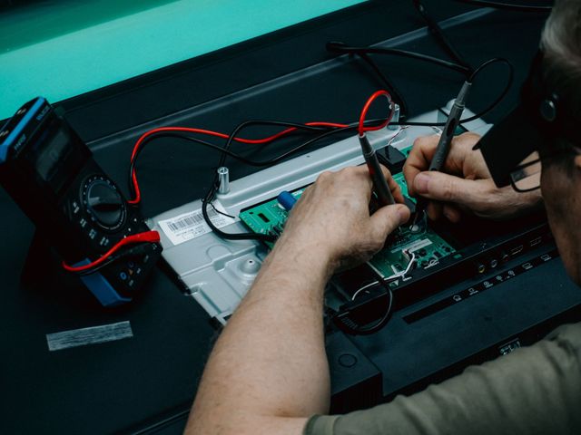 Manos reparando una placa de circuito impreso en un banco de trabajo con cables rojos y negros