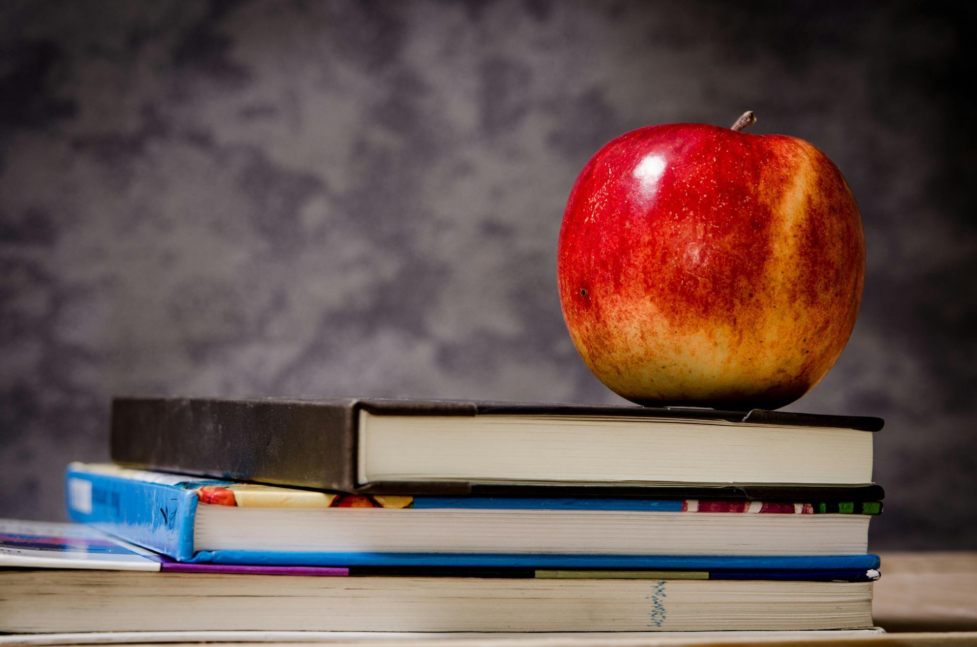 Red apple atop a stack of books, against a mottled gray backdrop.