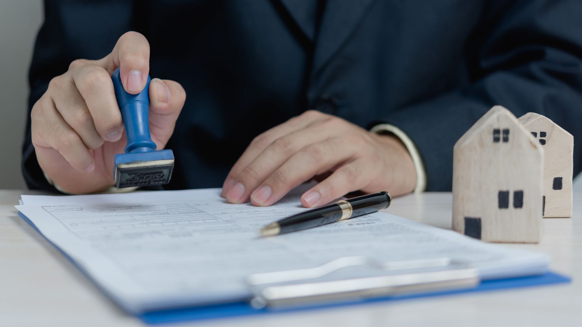 Hand stamping a document beside a pen and small wooden house model on a desk
