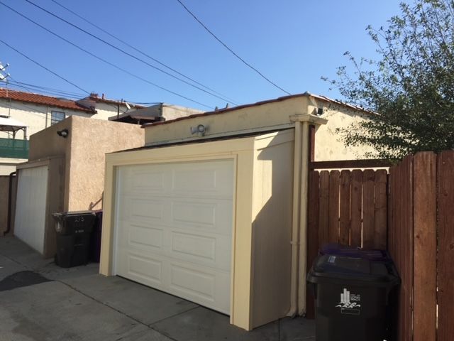 Three garages in alleyway, beige and tan buildings, wooden fence, black trash cans.