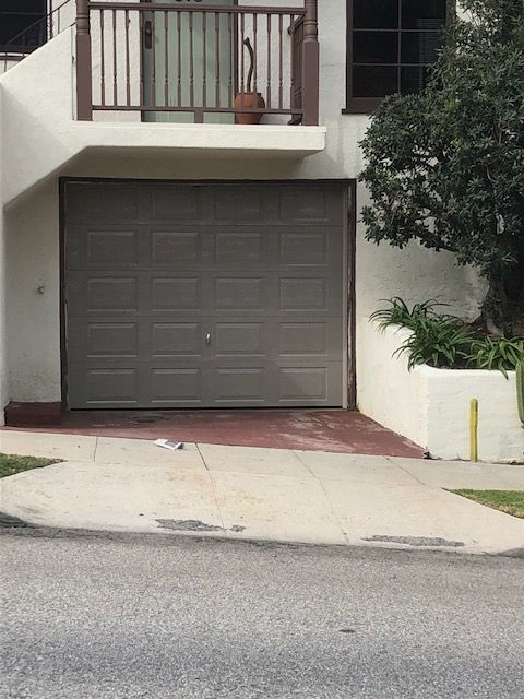 Brown garage door beneath a balcony. Gray driveway leads to a street.