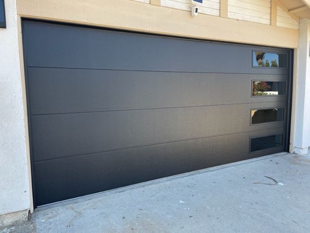 Black garage door with three windows on right side, set in a concrete driveway.