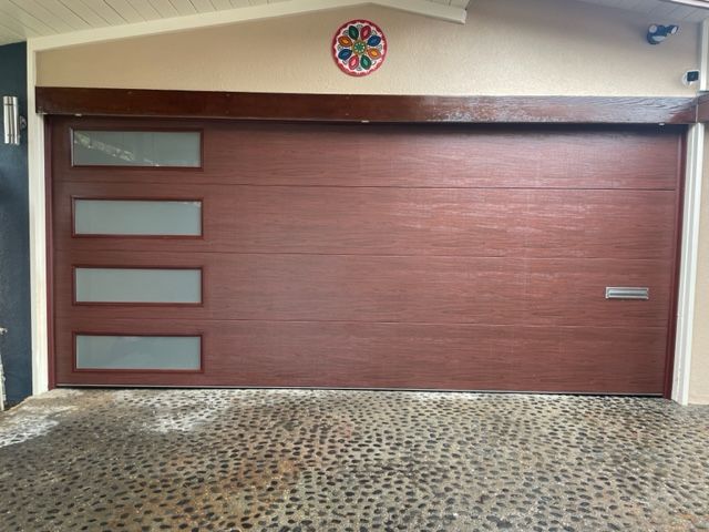Brown garage door with horizontal panels, three rectangular frosted windows, and a pebble driveway.