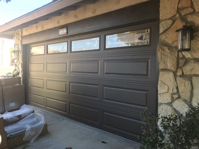 Brown garage door with rectangular windows above, next to a stone wall and a light fixture.