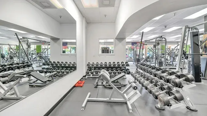 A brightly lit gym with rows of dumbbells, weight benches, and exercise machines.