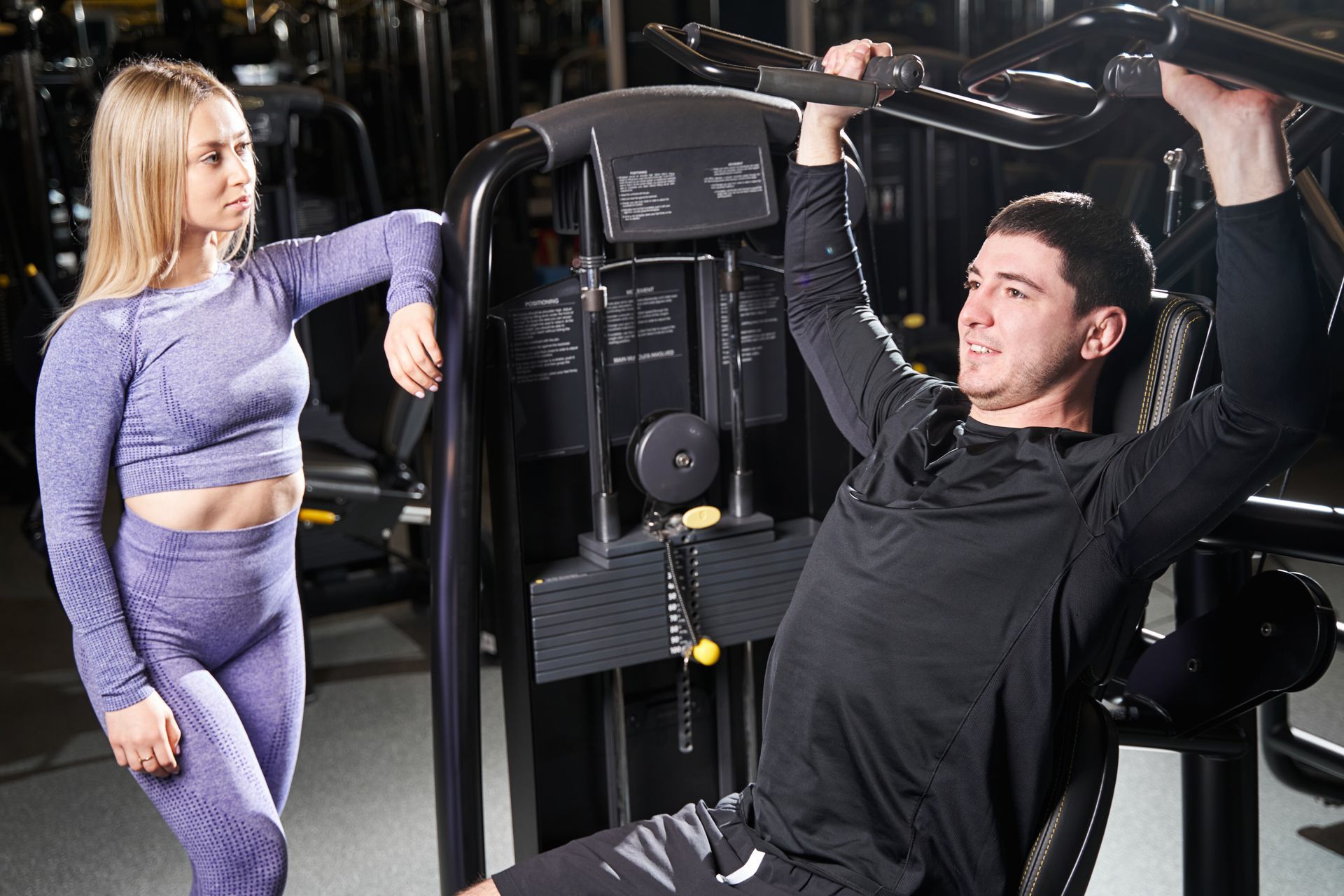 A woman is standing next to a man using a machine in a gym.