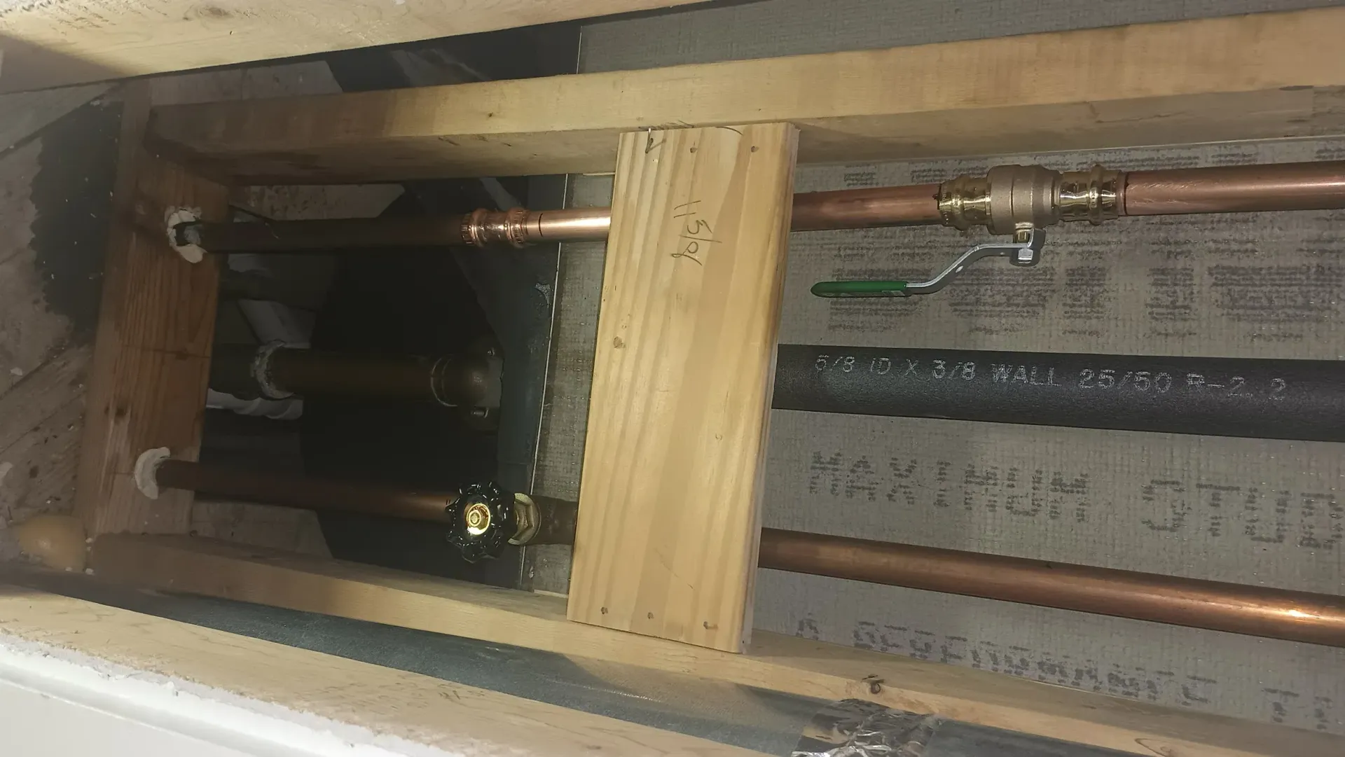 Plumbing access opening with copper pipes, valves, and insulation behind a wooden frame.