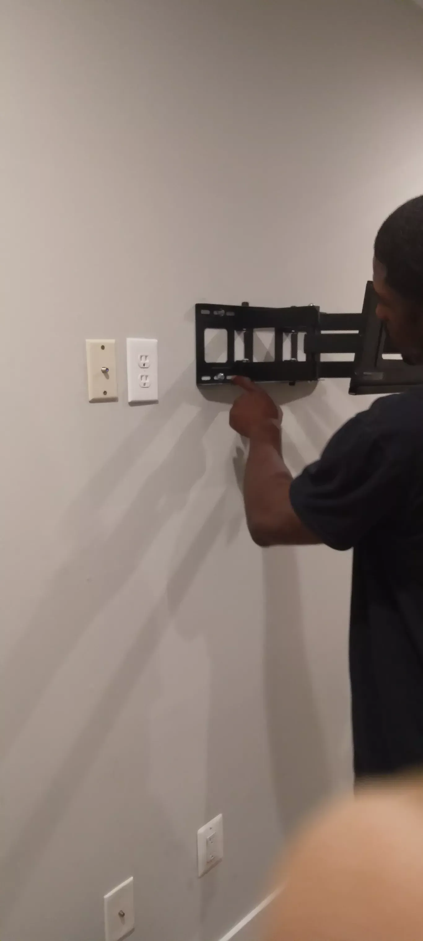 Person mounting a black wall bracket on a gray wall beside electrical outlets