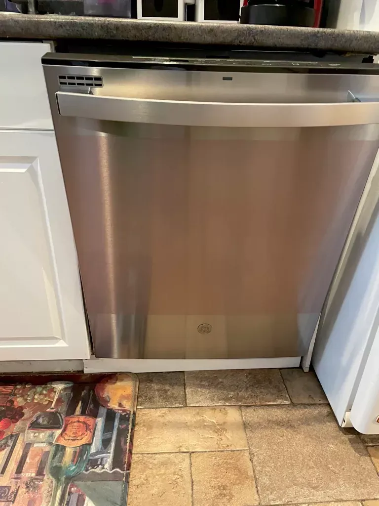 Stainless steel dishwasher beneath a kitchen countertop, flanked by white cabinets and tile flooring.