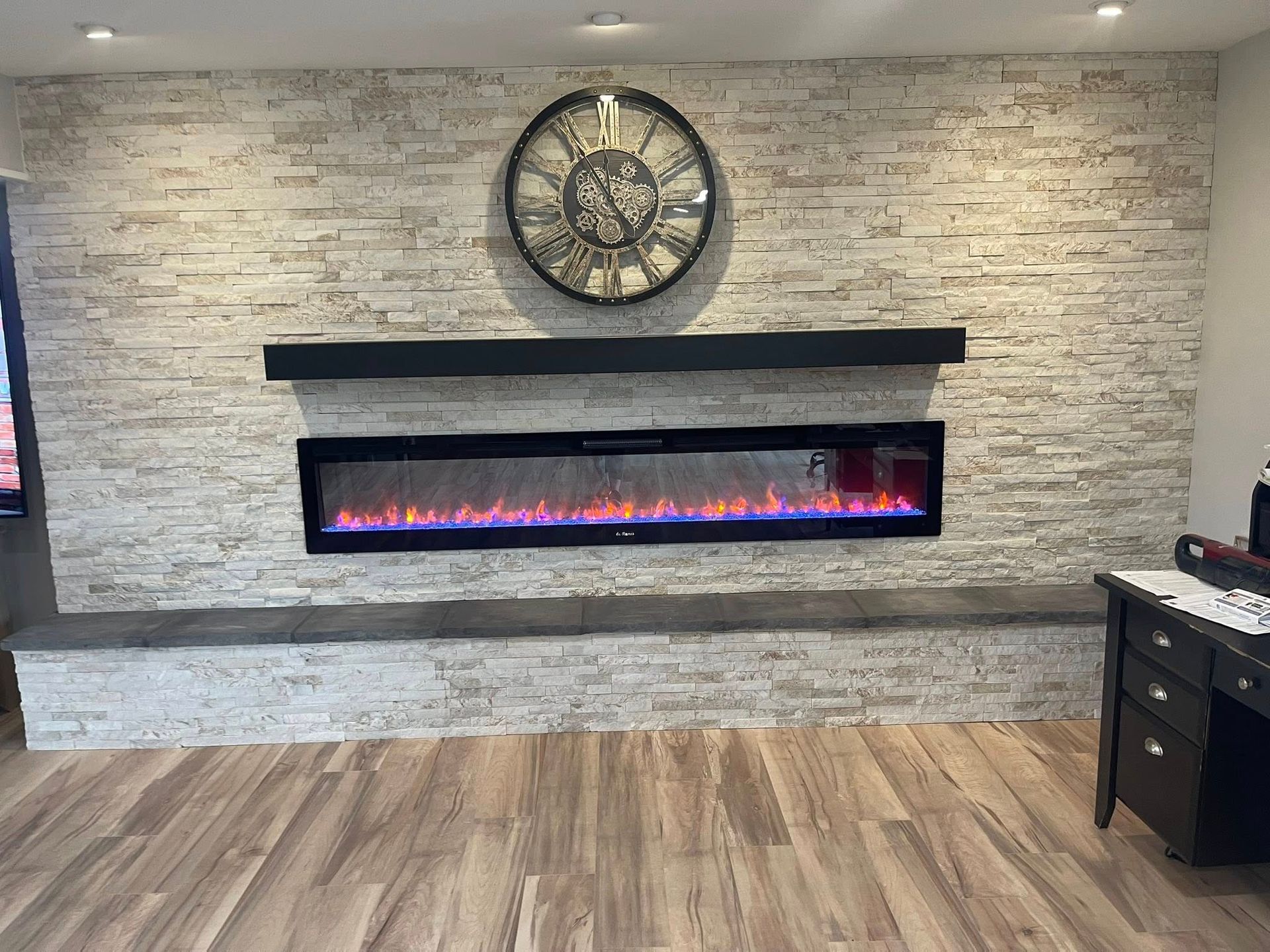 Fireplace with faux stone wall, black mantel, large clock, and wood flooring.