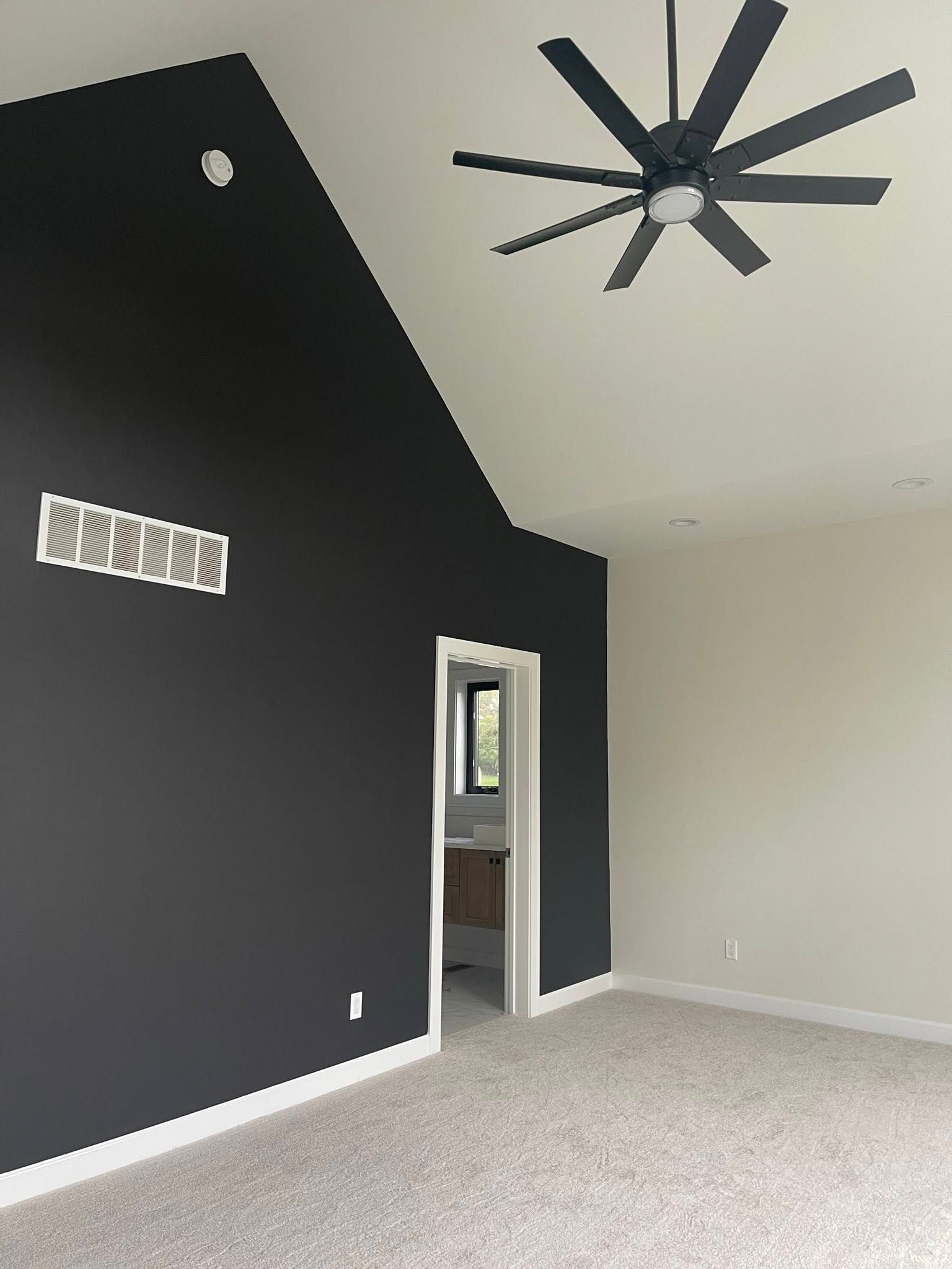 A modern bedroom with a black accent wall, white ceiling and fan, and neutral carpet. A doorway leads to a bathroom.