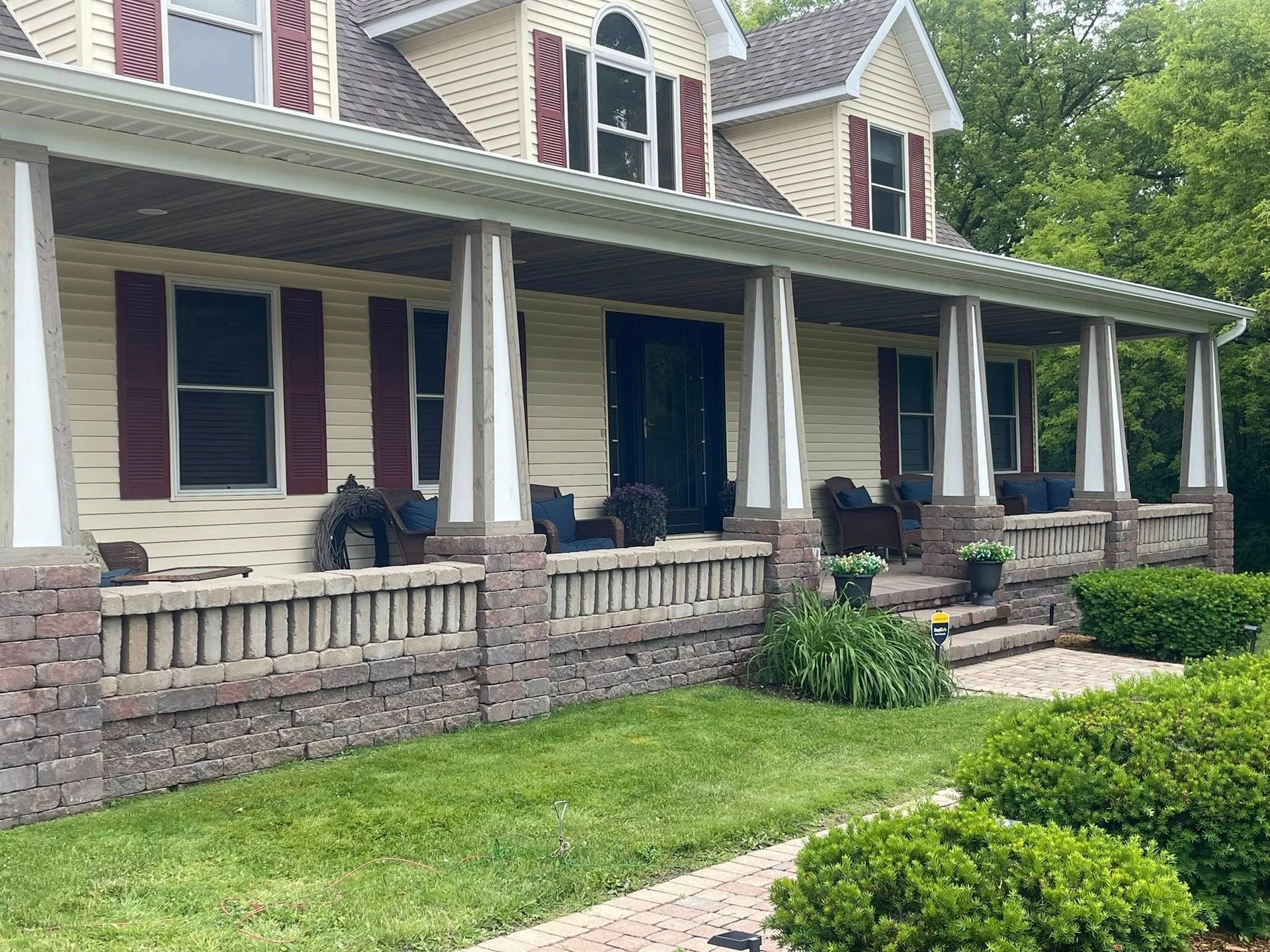 Beige house with porch, red shutters, and brick details. Green lawn and bushes in foreground.