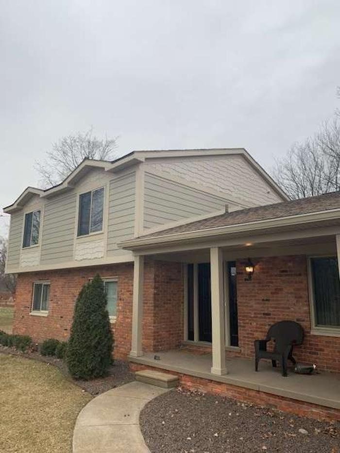 Two-story brick house with light gray siding. A covered porch has a chair. Cloudy day.