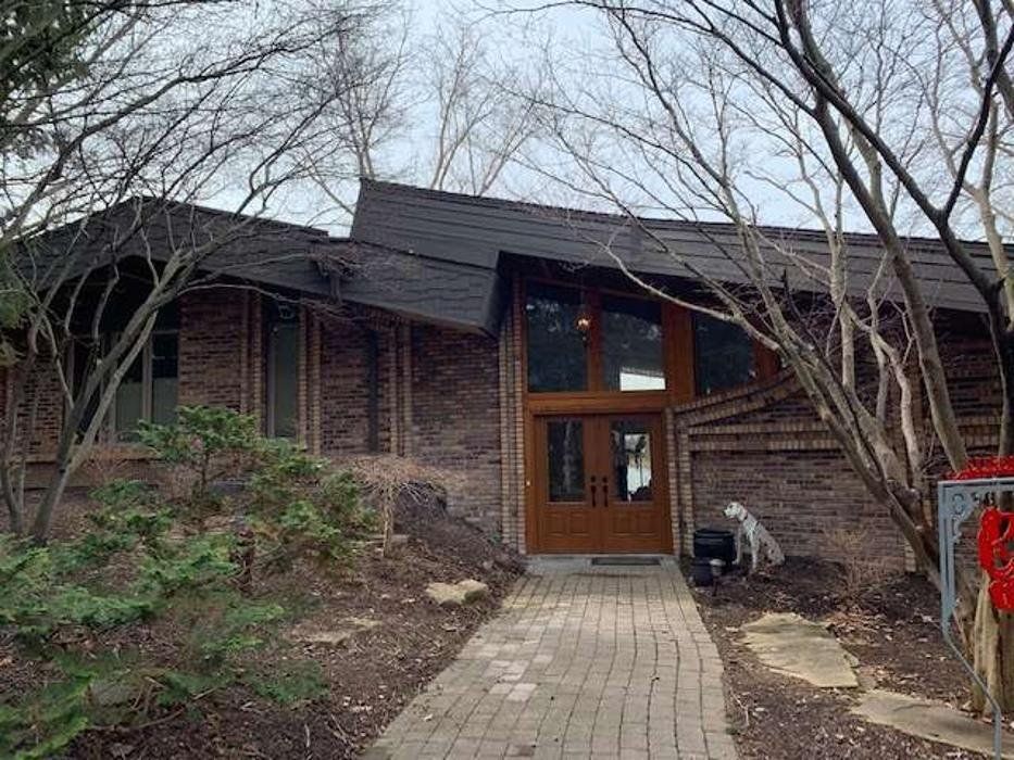 Brick house with a dark roof and large wooden front doors, approached by a brick path, surrounded by trees.