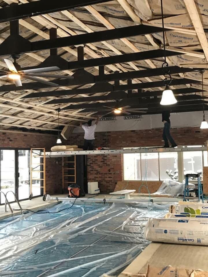 Construction inside a pool area. Two workers on scaffolding are working on the ceiling. Pool is covered in blue plastic.