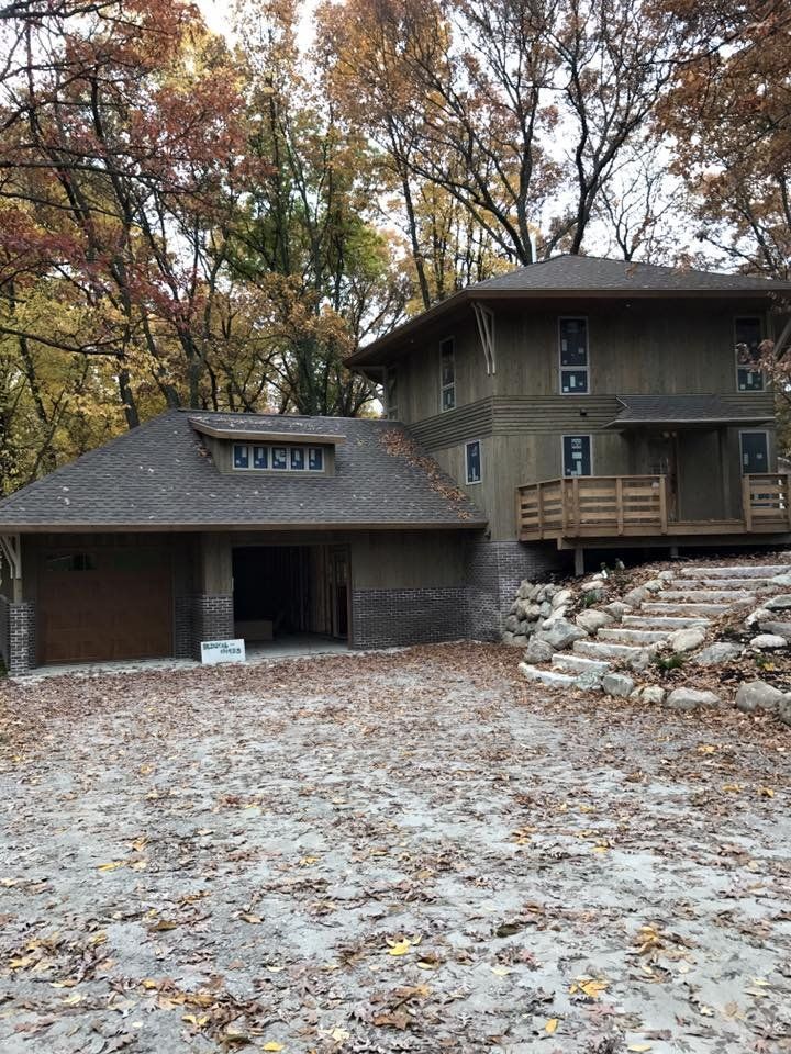 Two-story home with attached garage in a wooded area. Exterior is tan with brown roofing. Leaves cover the ground.