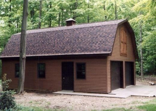 Brown barn-style garage with two bays, a door, and windows. Set in a wooded area with a concrete pad.