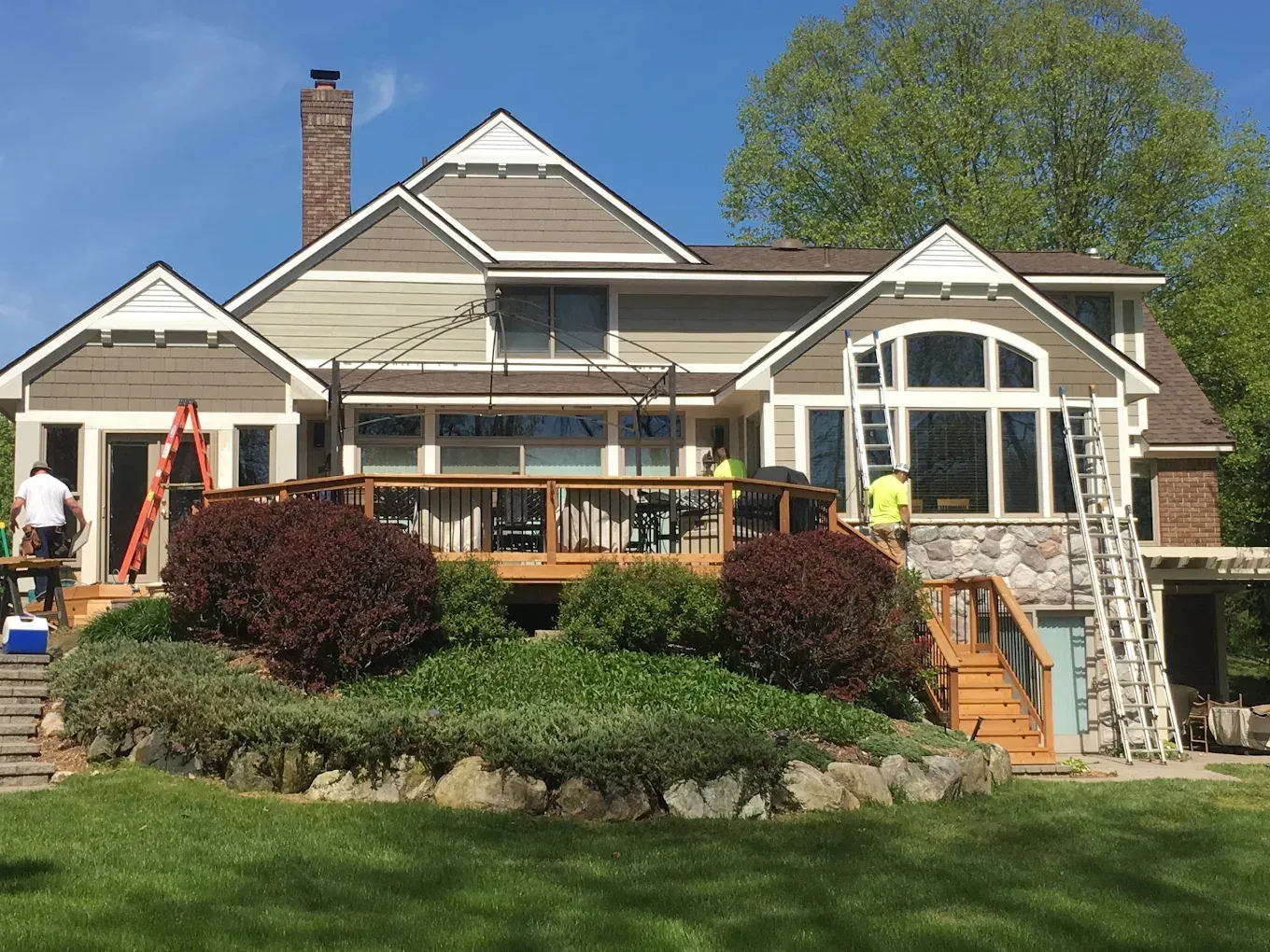 Two-story house with tan siding, wooden deck, and workers on ladders. Landscaping with bushes and rocks in yard.