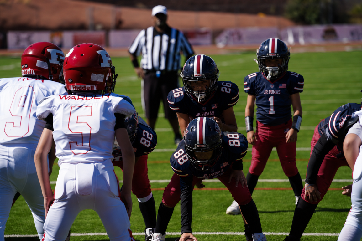 Football players in action on a grassy field; one runs with the ball, pursued by two opponents.