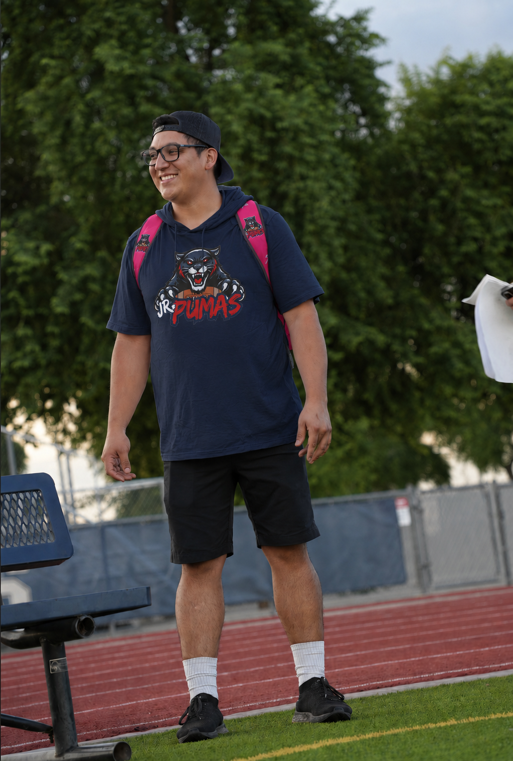Man with crossed arms on a football field. He is wearing a blue polo shirt, khaki pants, and has a serious expression.